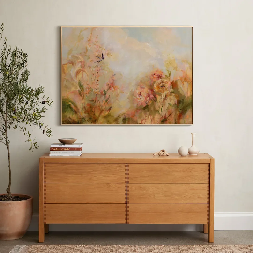 A wooden sideboard with decorative objects on top, a large floral artwork hanging above, an olive tree in a pot to the left, and a beige rug in front.