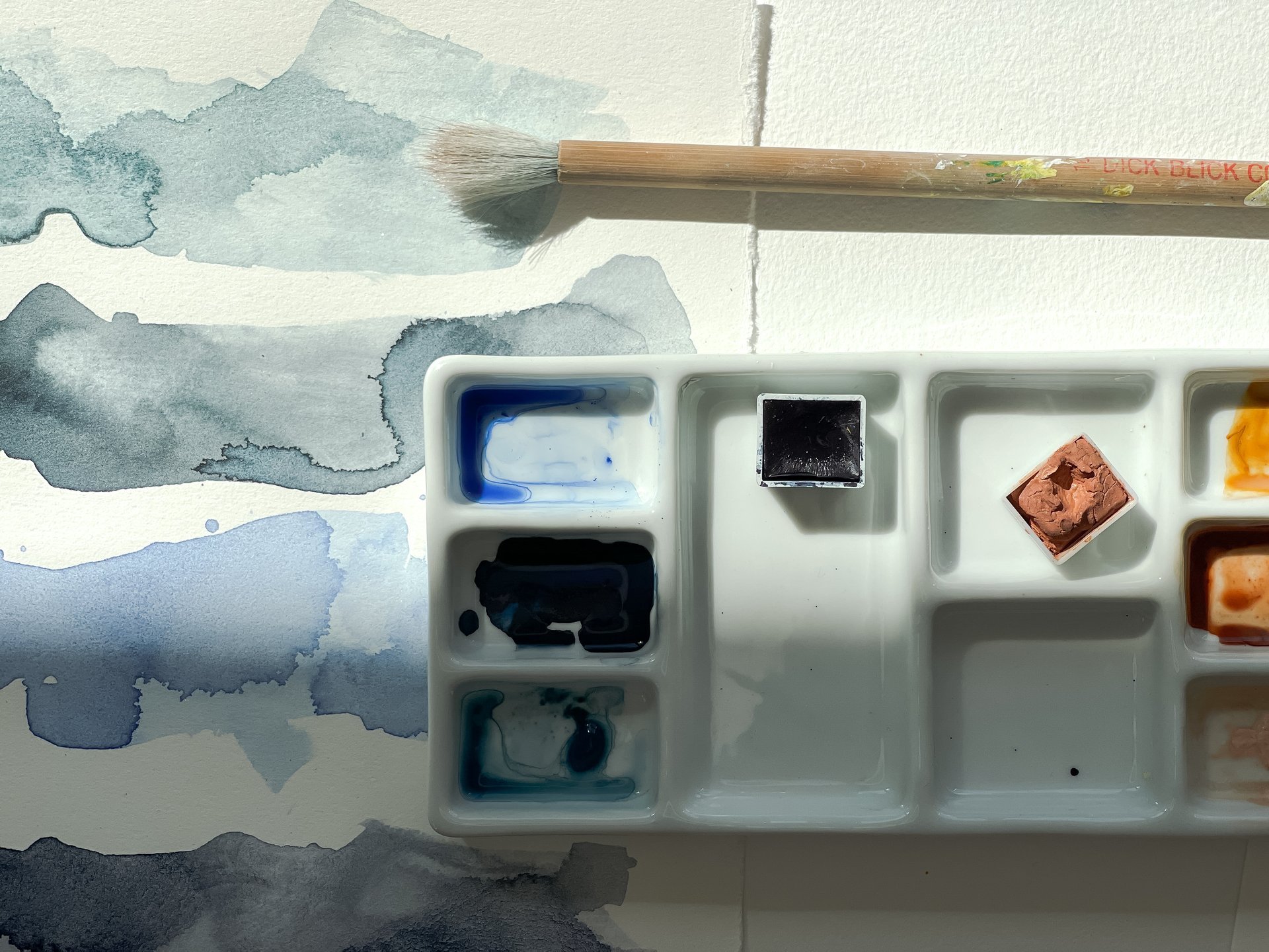 Watercolor paints and palette on a white surface, with blue watercolor splashes and a paintbrush.