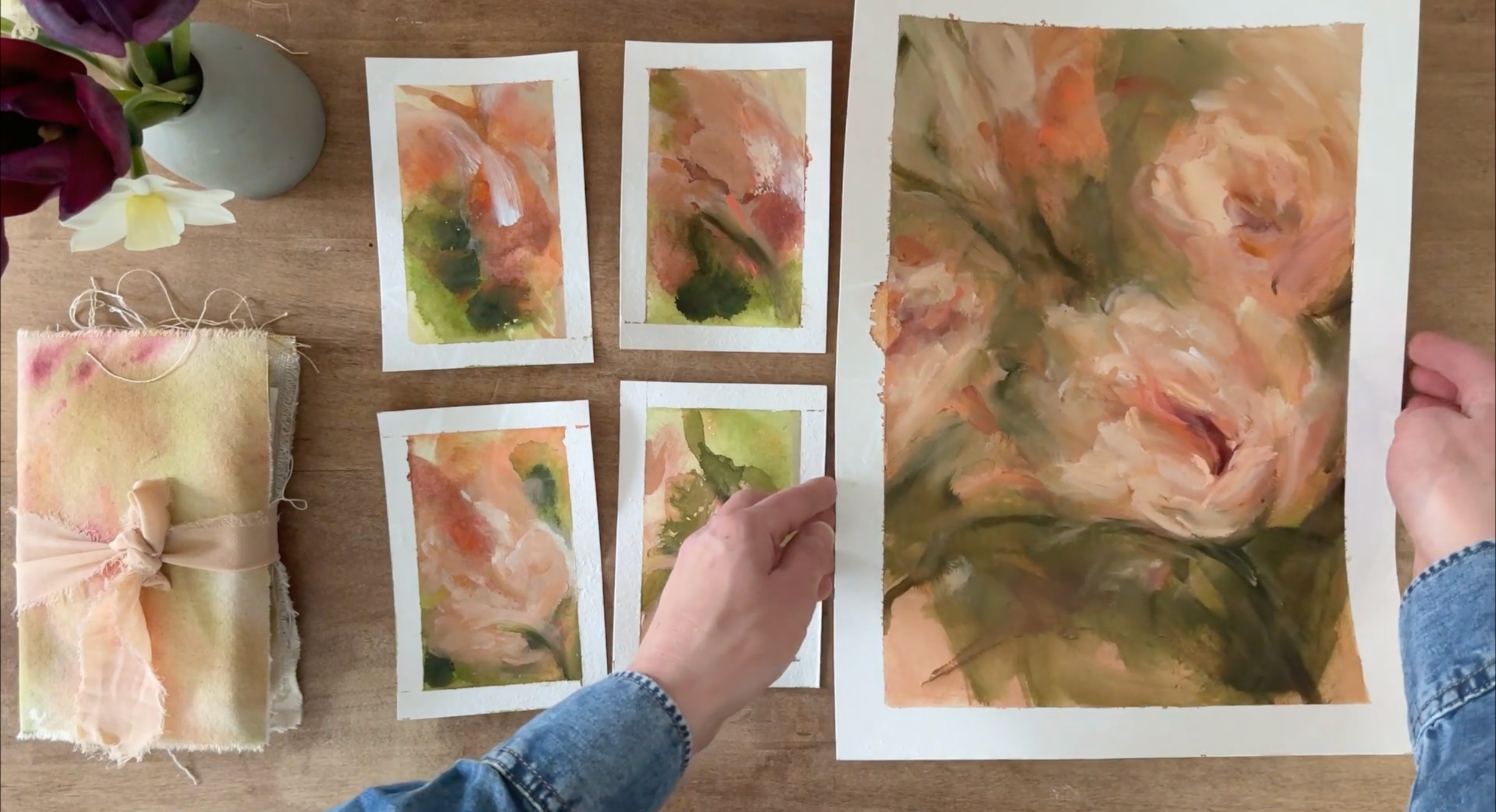 Person holding a large abstract floral watercolor painting with multiple smaller watercolor pieces on a wooden table, along with a bundle of fabric and a vase with flowers.
