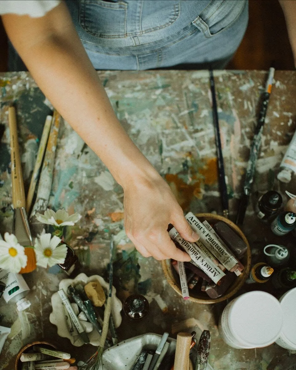 A person is arranging or selecting art supplies on a splattered, paint-covered table. The table has brushes, markers, small bottles of paint, and a basket with newspaper headlines.