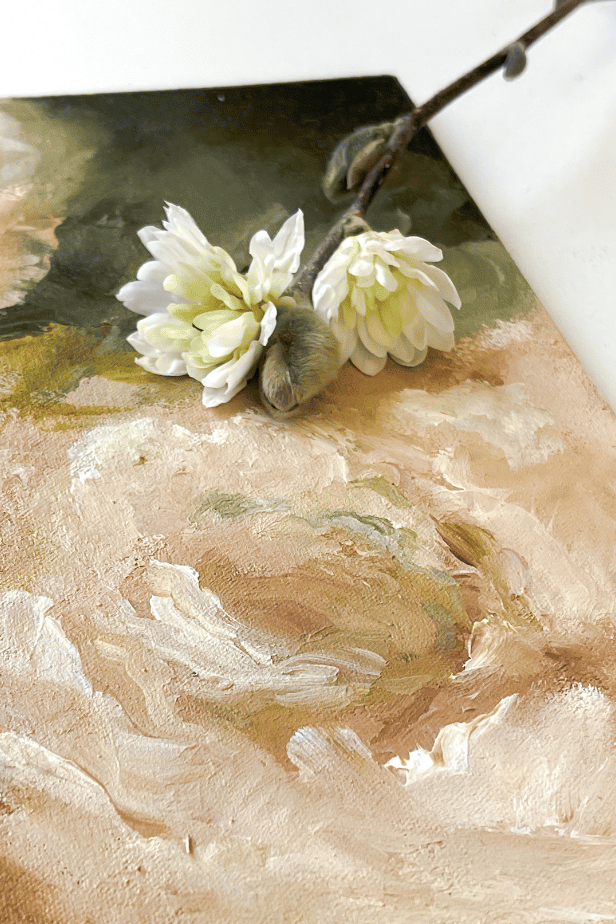 A close-up of two white flowers with yellow centers on a brown branch, lying on a textured, abstract painting with earth tones.