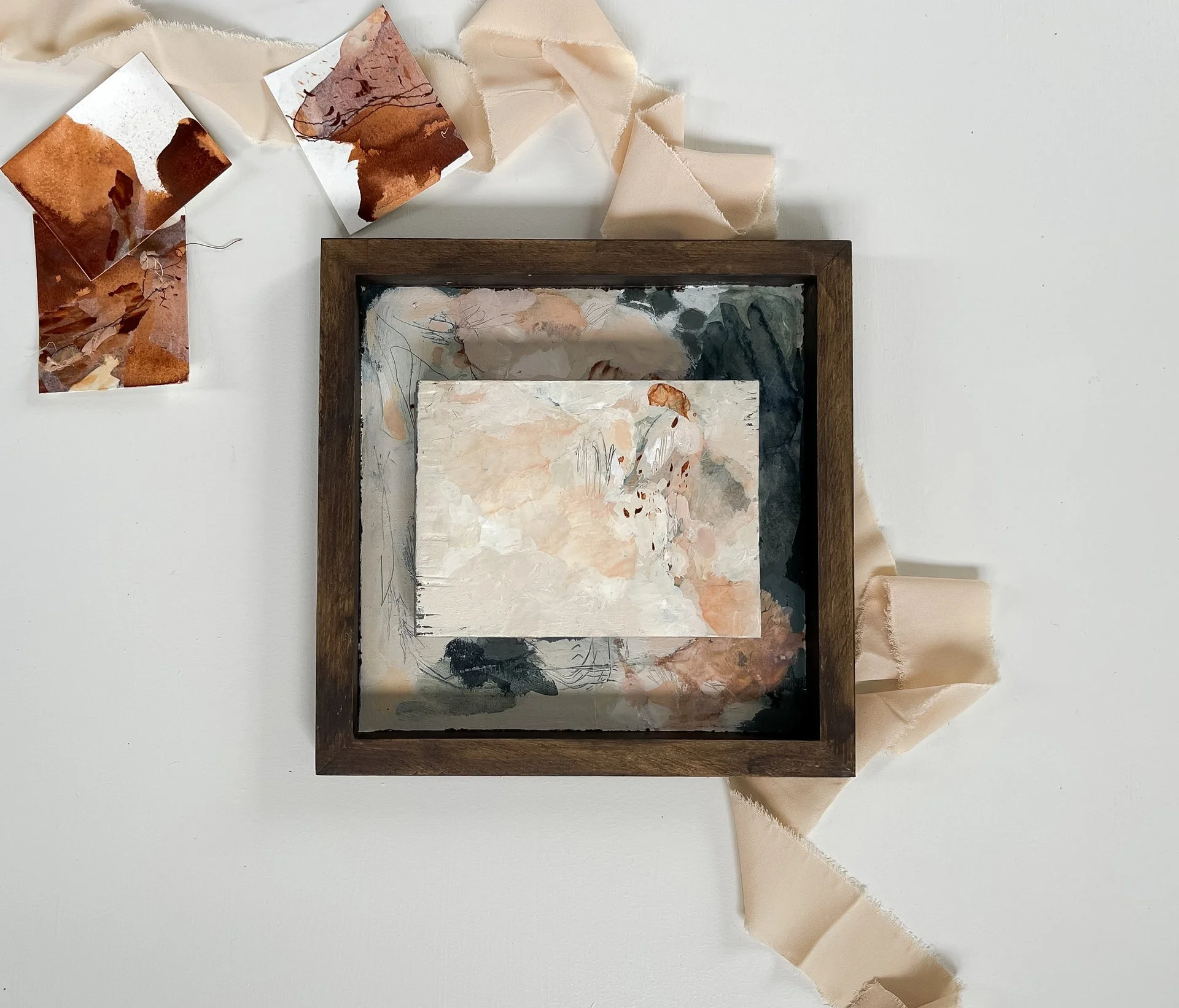 Abstract watercolor painting with shades of beige, white, gray, and brown inside a wooden frame, surrounded by torn beige paper strips and a torn paper rectangle on a white surface.