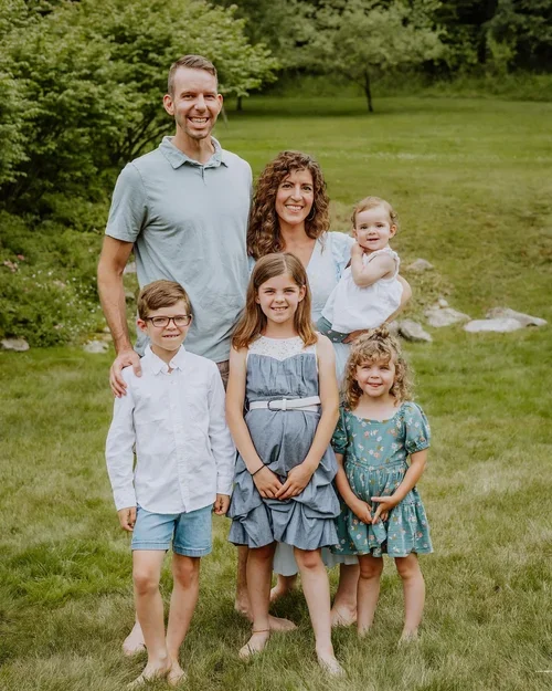 A family of six standing outdoors on a grassy area, smiling at the camera. The family consists of two adults and four children, with trees in the background.