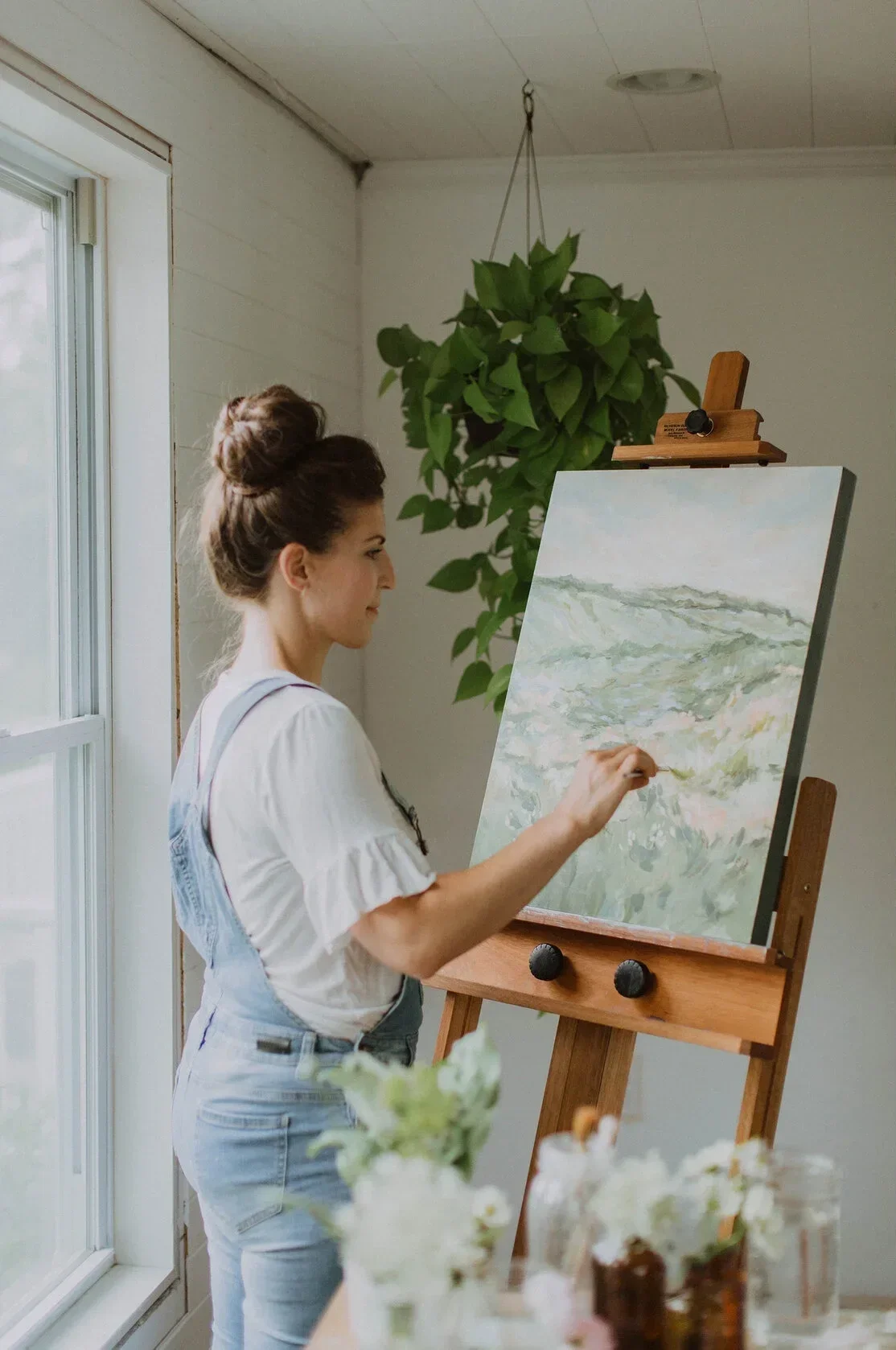 A woman painting an landscape on a canvas set on an easel in a bright art studio.