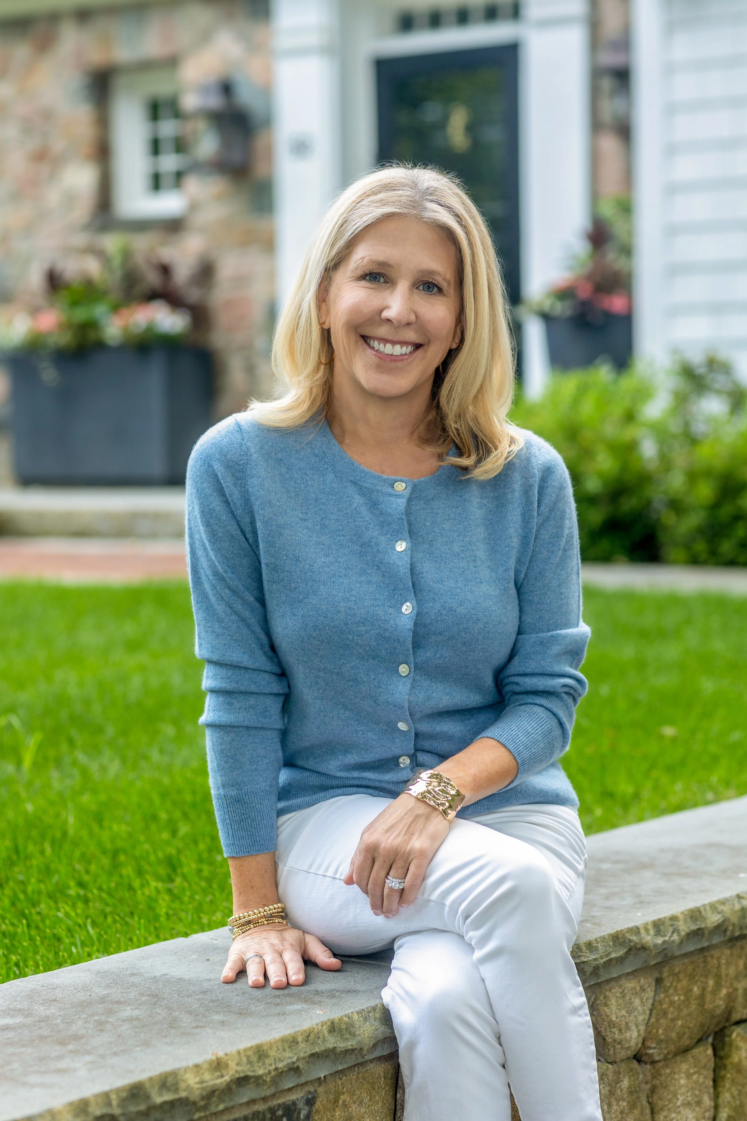 A woman with blonde hair smiling, sitting on a stone bench outdoors, wearing a light blue cardigan and white pants, with a house and greenery in the background.