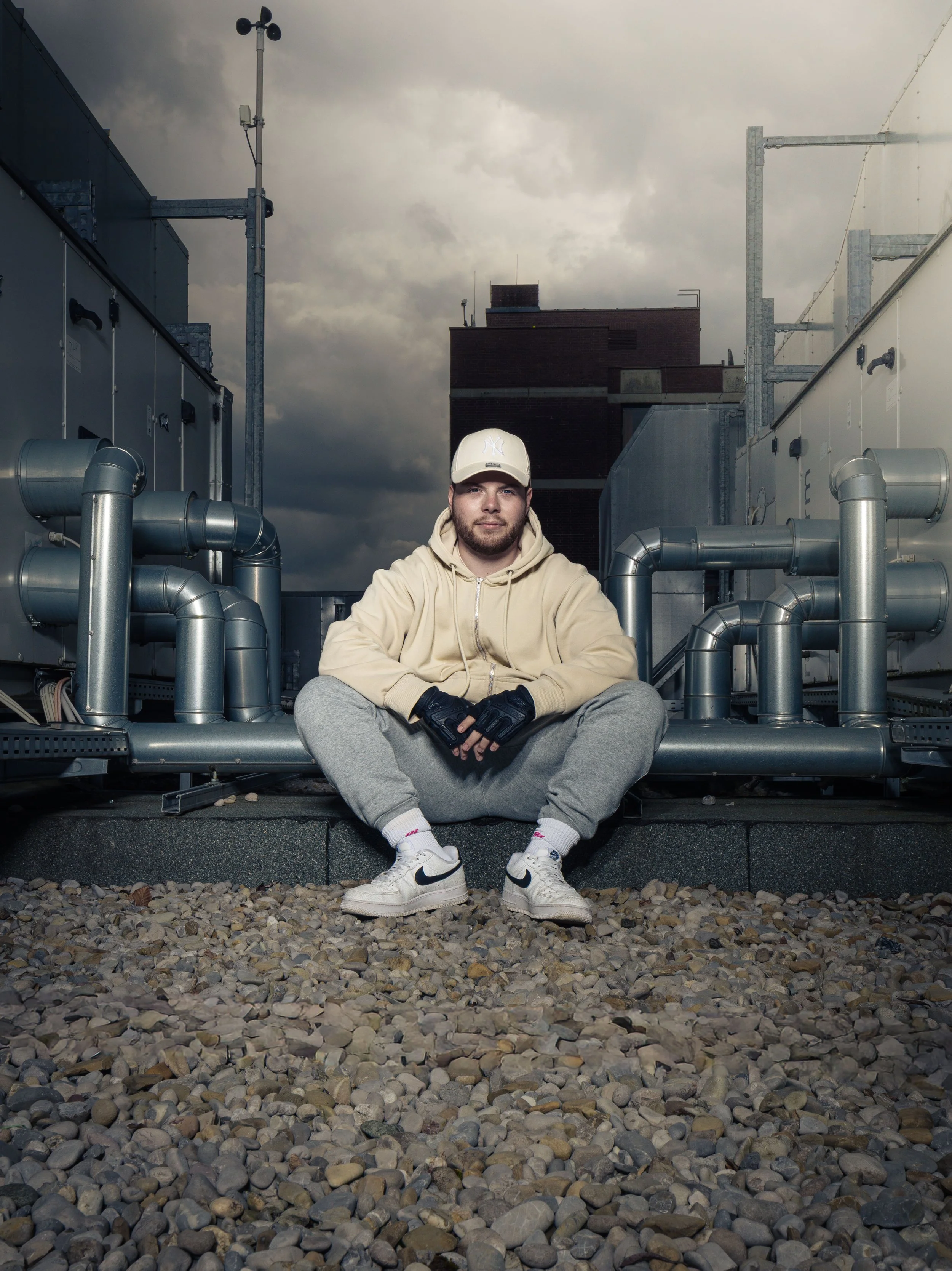 Junge Mann sitzt auf einem Dach mit Rohren und technischen Anlagen, trägt graue Jogginghose, beige Hoodie, weiße Nike Schuhe, weiße Cap, schwarze Handschuhe, vor dunklem Himmel mit Wolken.