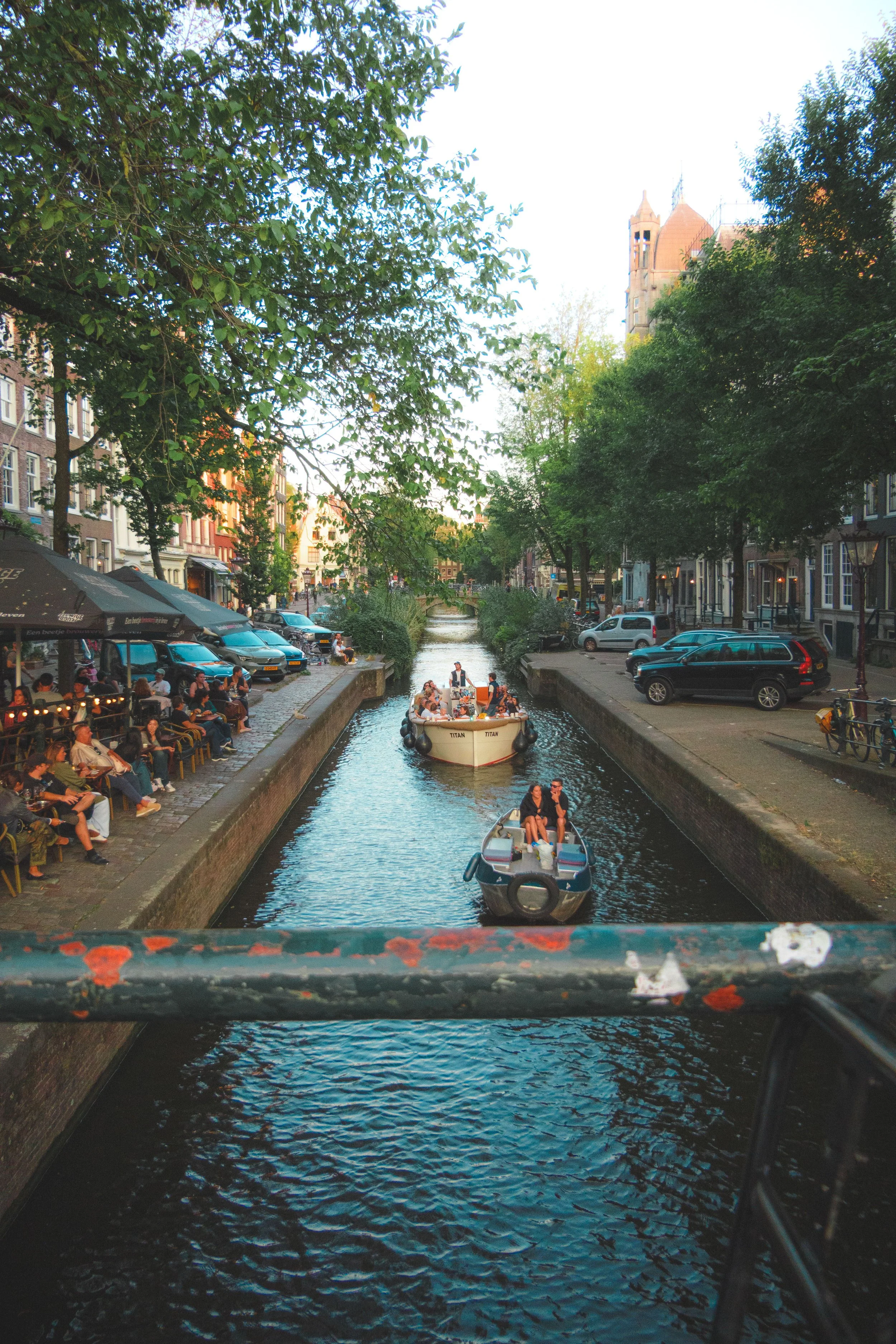 Touristenboote fahren durch eine schmale Amsterdamer Gracht, gesäumt von Cafés und Bäumen