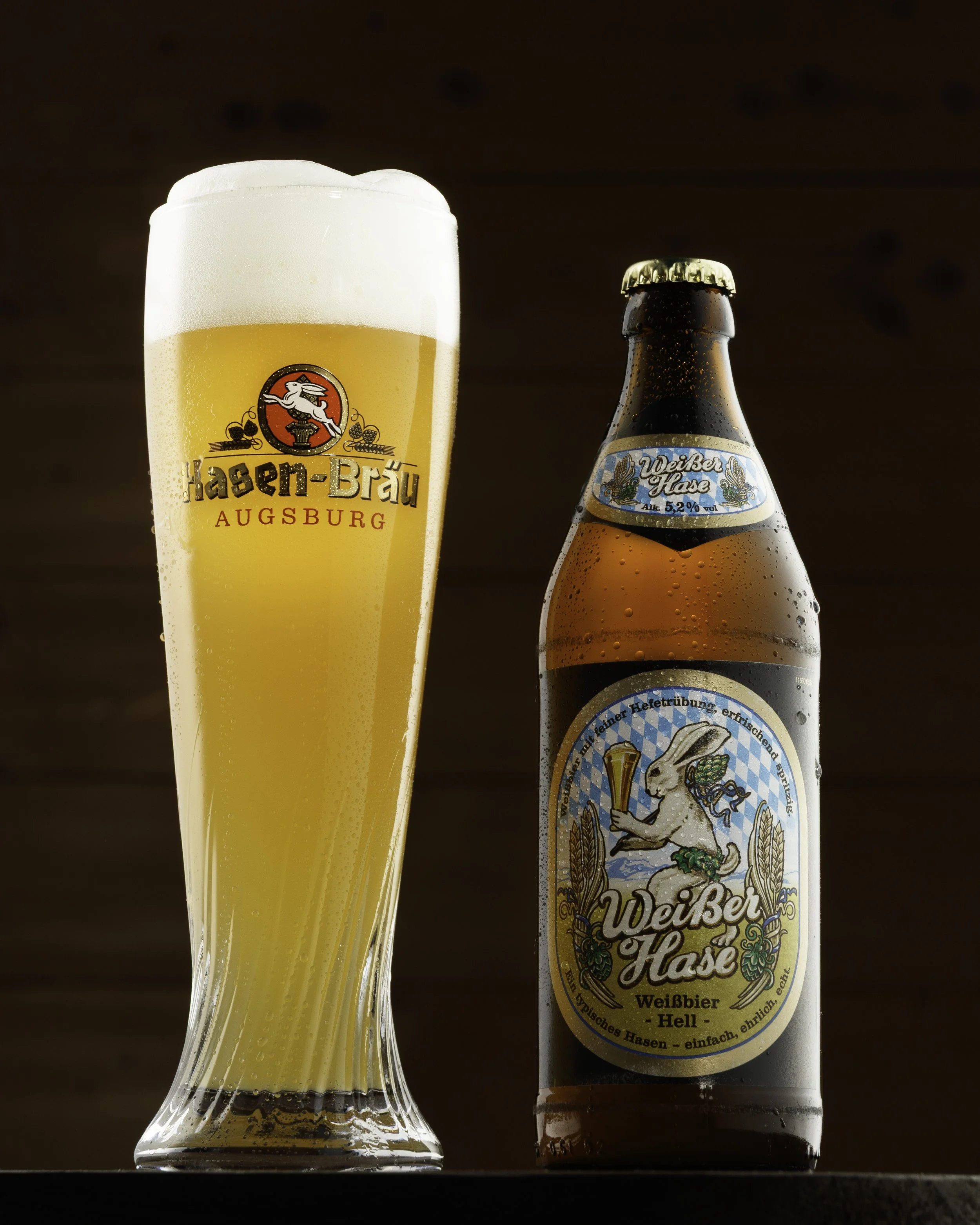Ein Bierglas mit hellem Bier und Schaumkrone neben einer Flasche Weizenbier mit bayerischem Etikett, beide auf einem dunklen Untergrund.