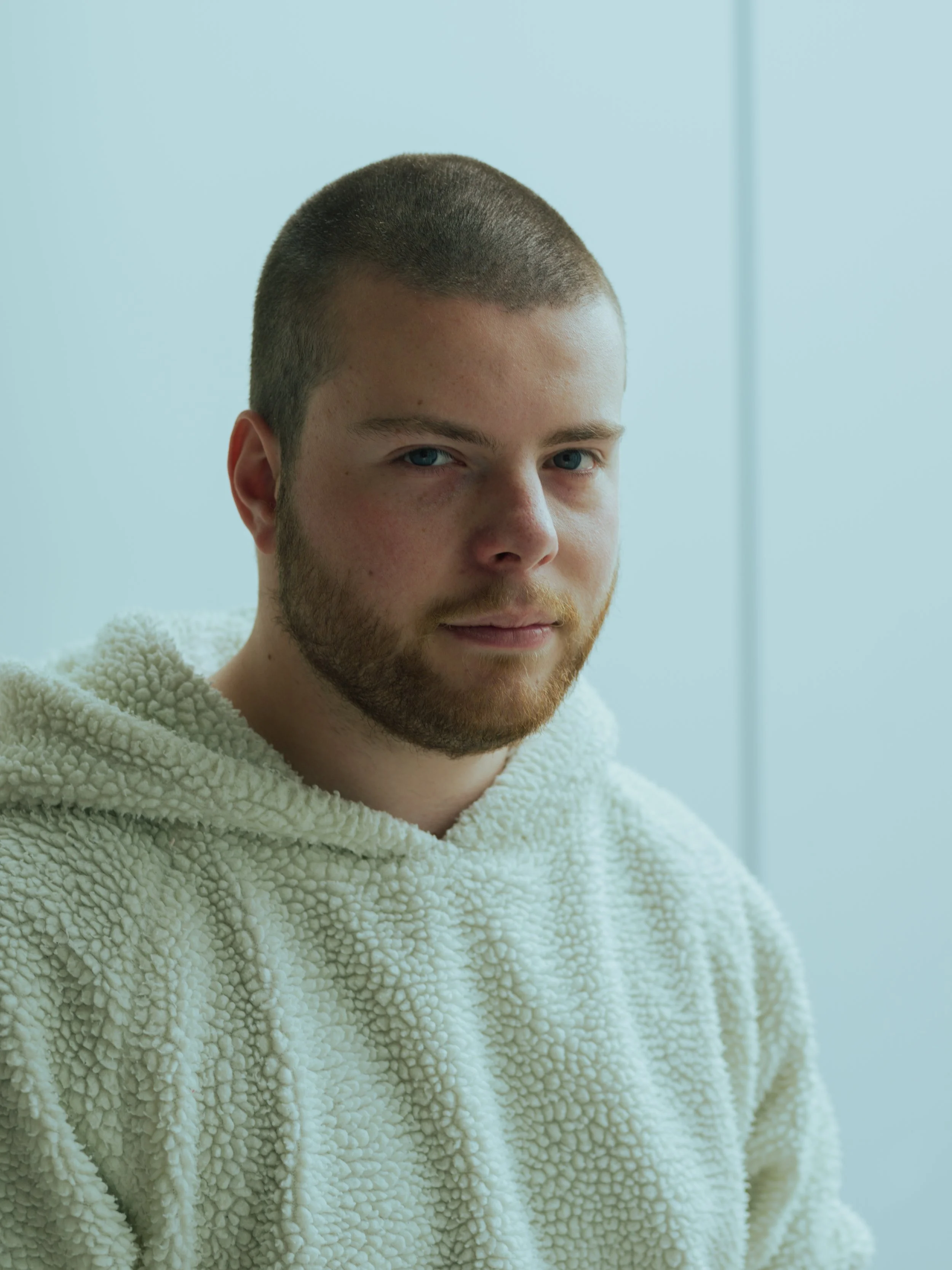 Junge Mann mit Kurzhaarschnitt und Bart trägt einen cremefarbenen, pelzigen Pullover vor einem hellblauen Hintergrund.