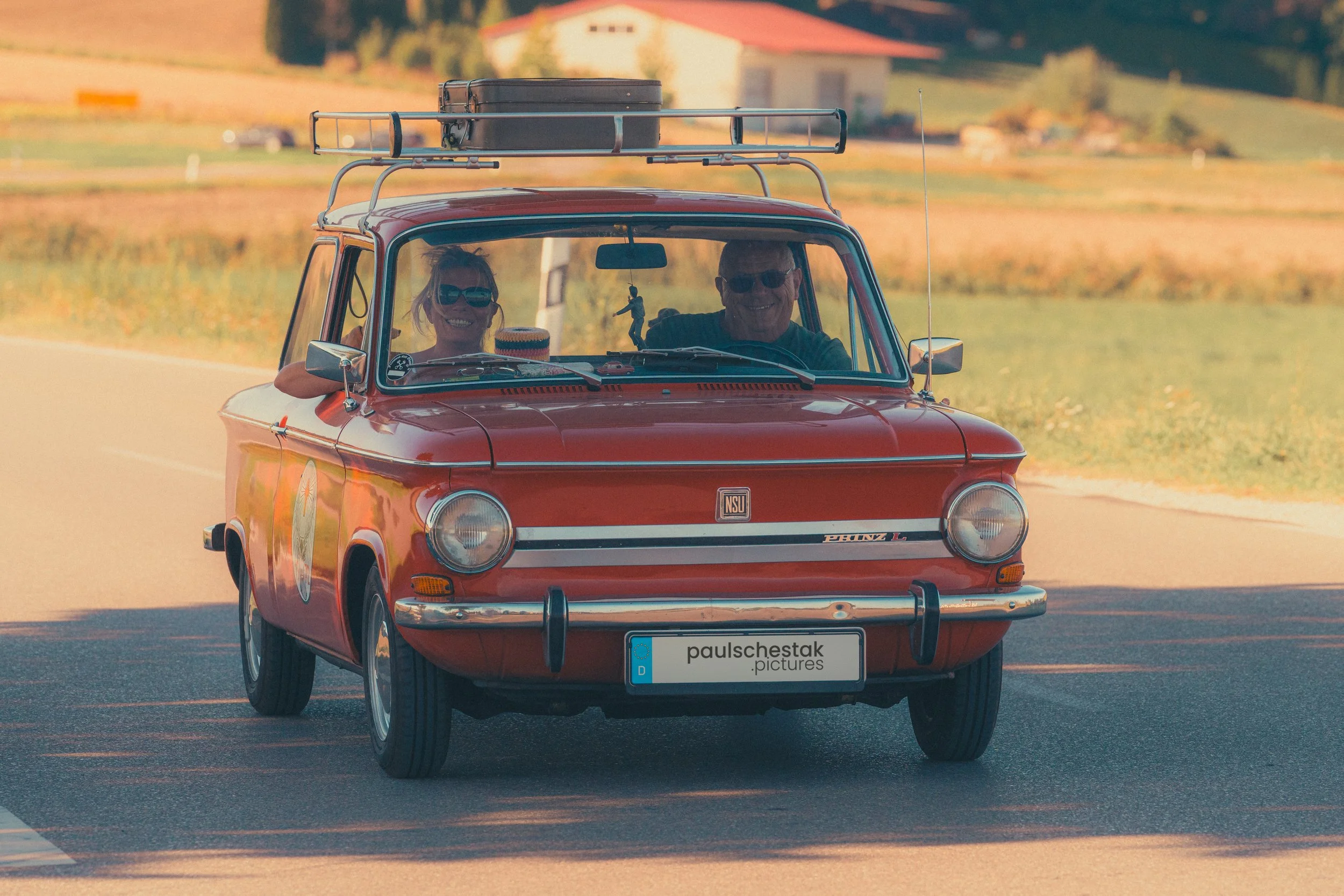 Roter Oldtimer-Kleinwagen mit Dachgepäckträger und zwei Insassen in Fahrt