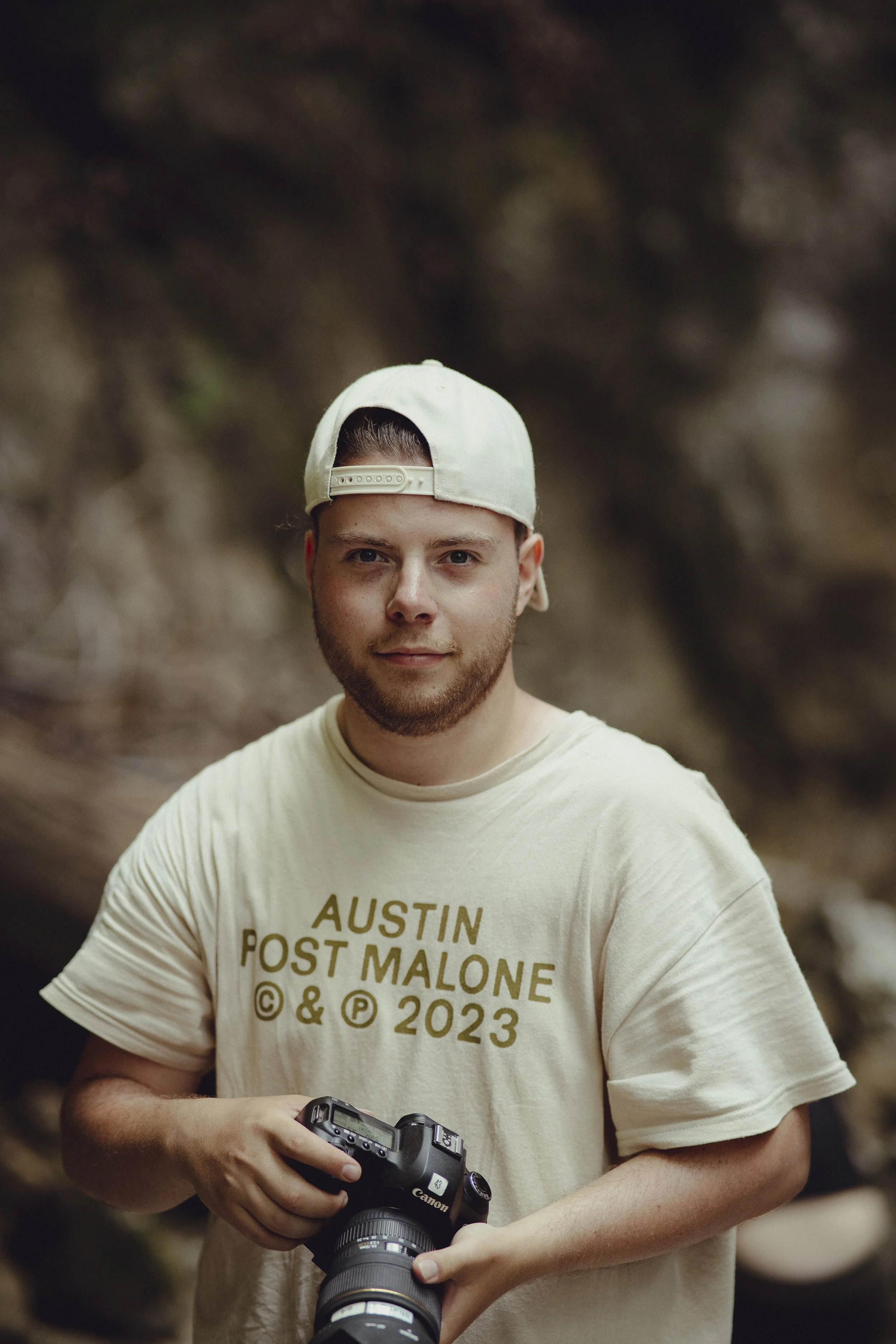 Junger Mann mit beige Baseballcap und T-Shirt, das 'Austin Post Malone' trägt, hält eine Kamera in den Händen und steht vor unscharfem Naturhintergrund.