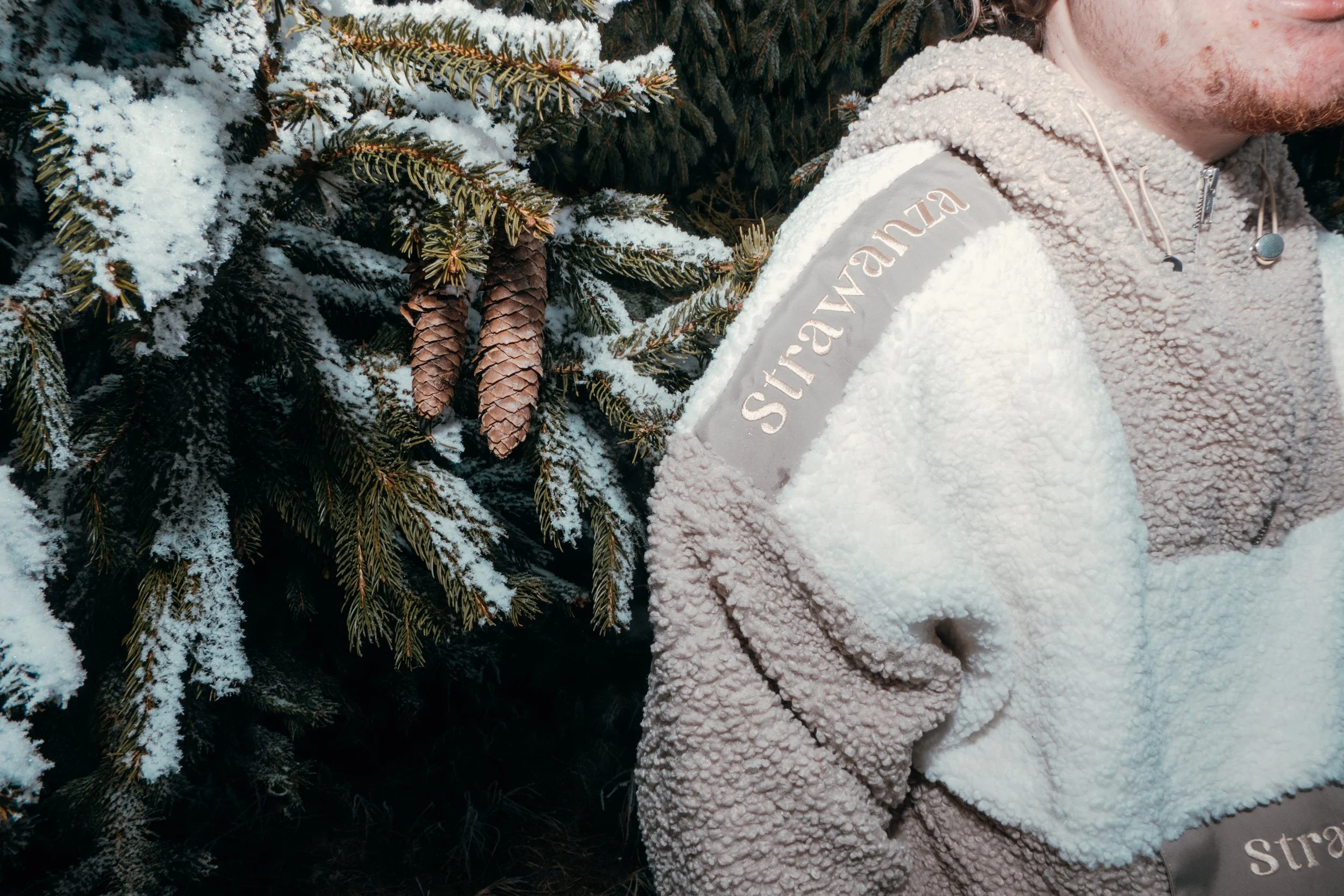 Ein Person trägt einen weißen, flauschigen Jacke mit einem Namensaufnäher mit dem Text 'Strawanza', neben einer Tanne mit Schneemanniveau. Die Schneetextur ist deutlich sichtbar, und die Person trägt auch Ohrhörer.