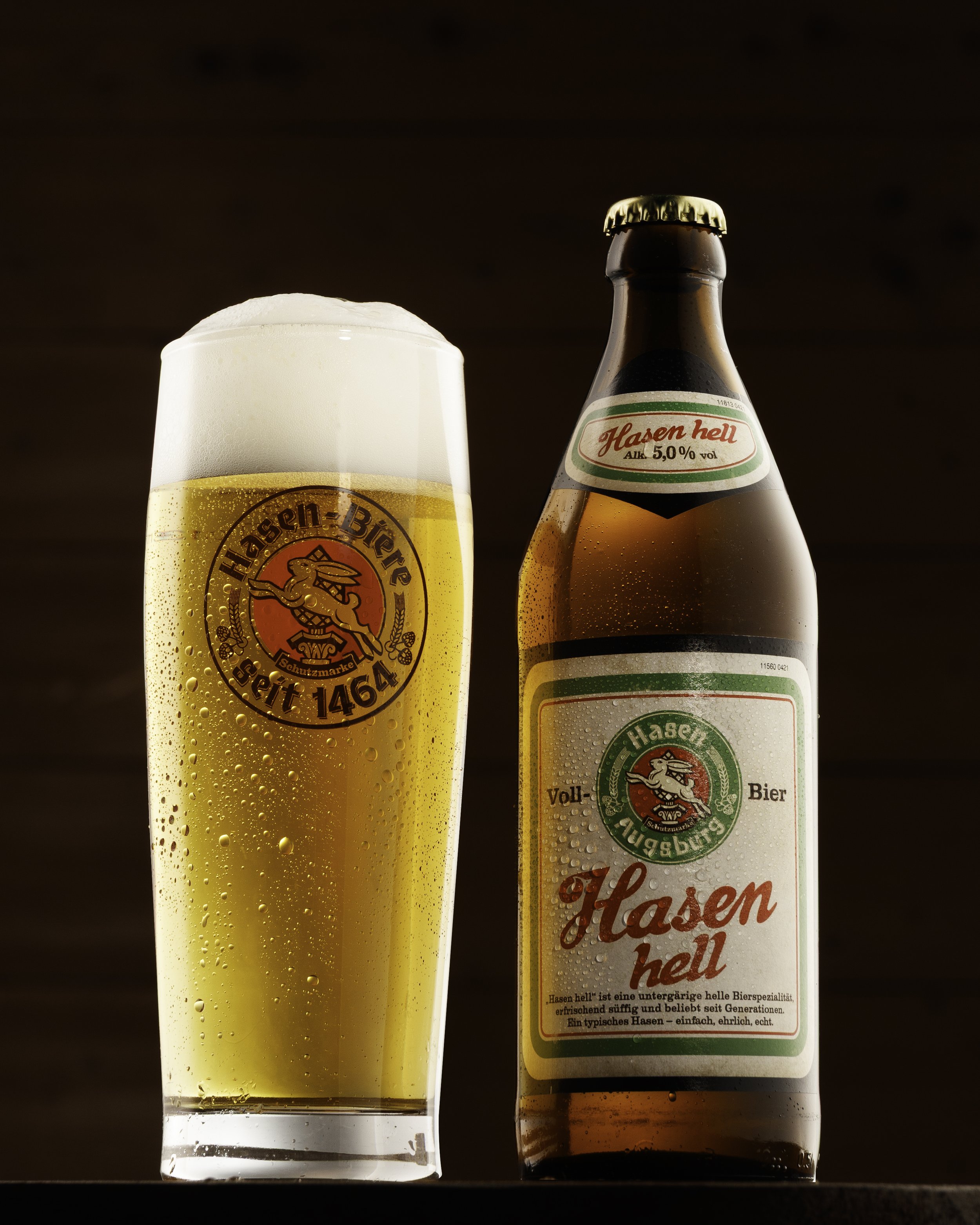 Ein Glas Bier mit Schaum neben einer Flasche Hasen hell Bier. Das Glas hat ein Emblem mit einem Hasenkopf und der Aufschrift 'Hasen Brau'. Die Flasche trägt ein Etikett mit der Aufschrift 'Hasen hell' und enthält 5,0 % Alkohol.
