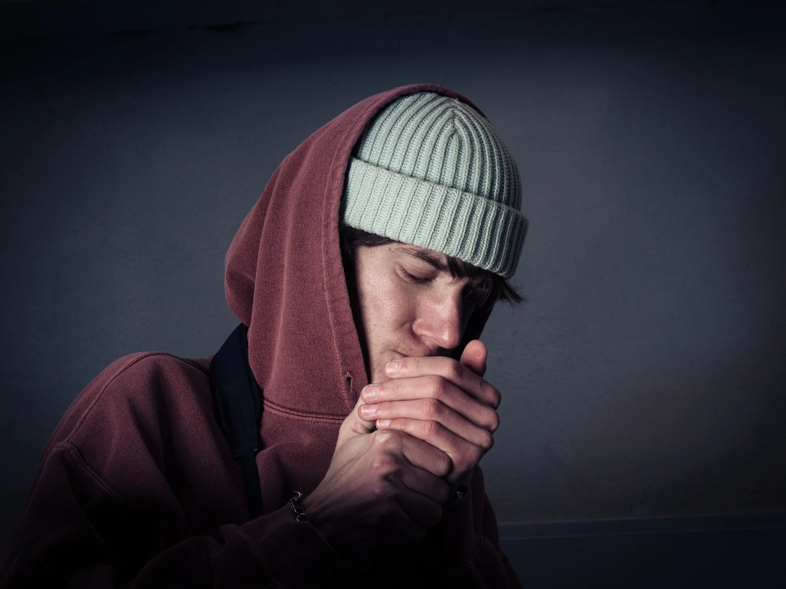 Junge Person betet mit zusammengelegten Händen, trägt grauen gestrickten Hut, rote Kapuzenjacke und hat die Augen geschlossen.