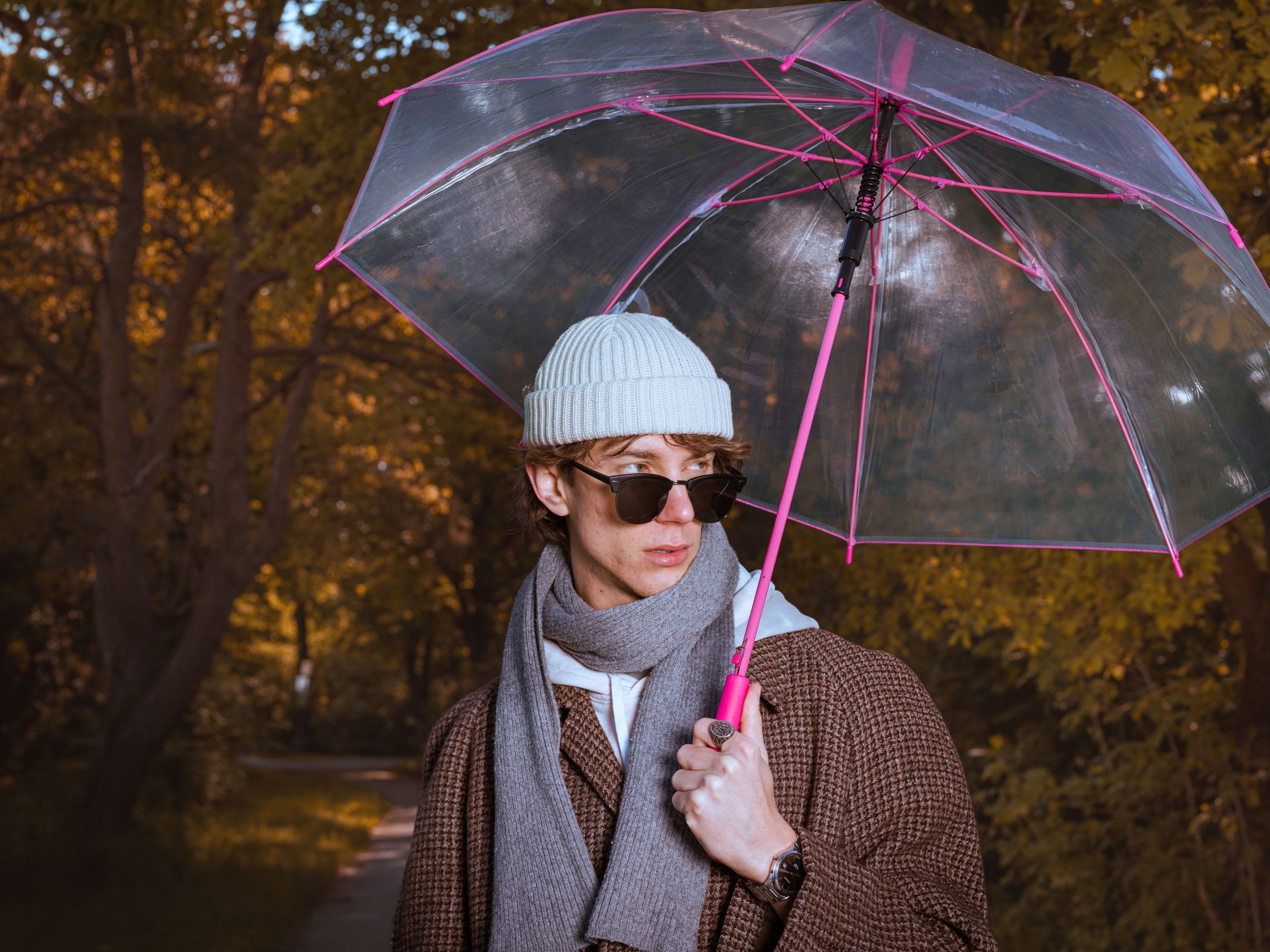 Junge Person mit Sonnenbrille, grauem Hut, braunem Mantel, grauem Schal, hält einen transparenten Regenschirm mit pinken Stangen bei Dämmerung im Park mit Herbstlaub im Hintergrund.