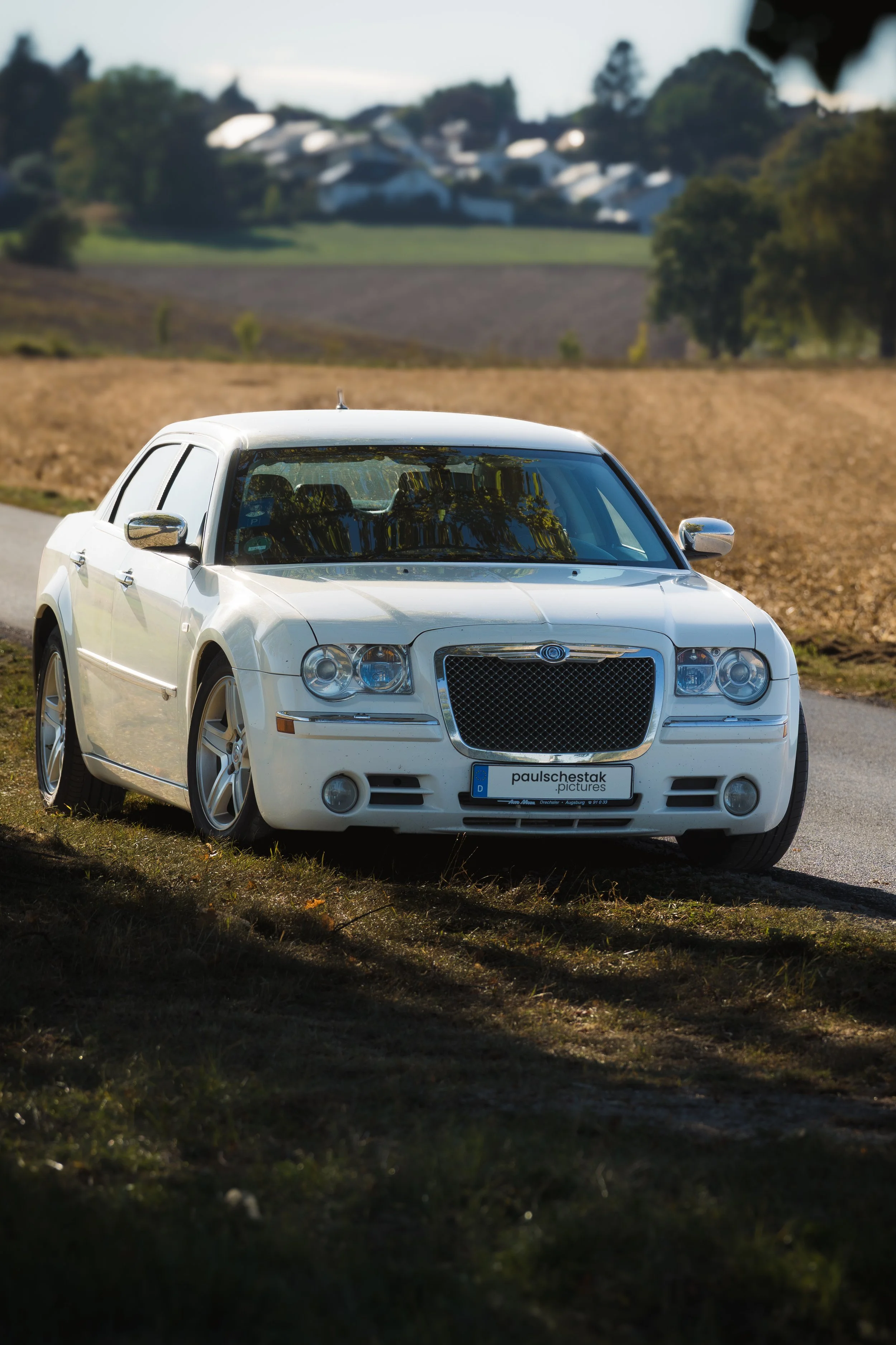 Weißer Chrysler 300C am Feldrand, im Hintergrund ein Dorf