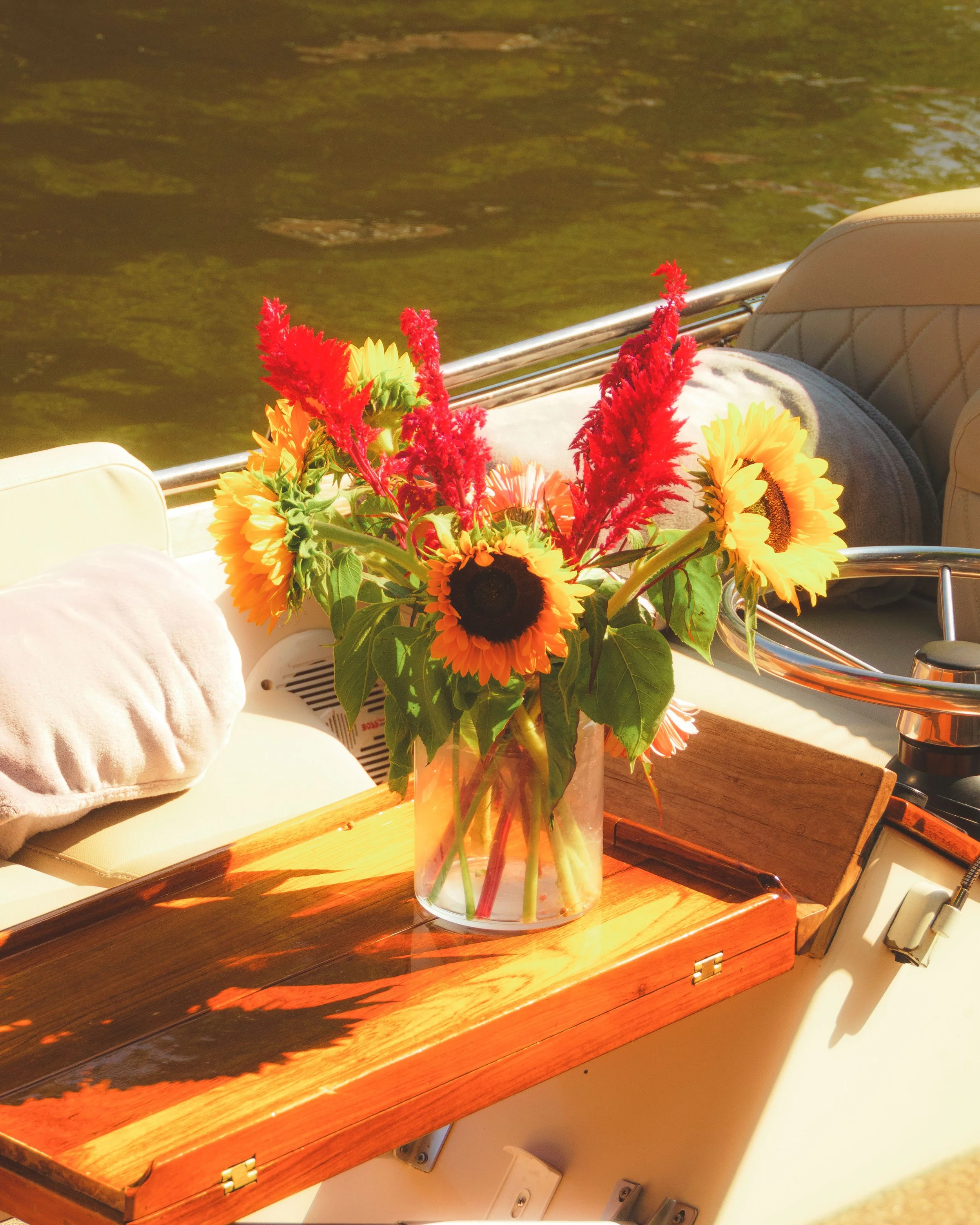 Bunter Blumenstrauß in einer Glasvase auf einem Bootstisch, im Hintergrund grünes Wasser