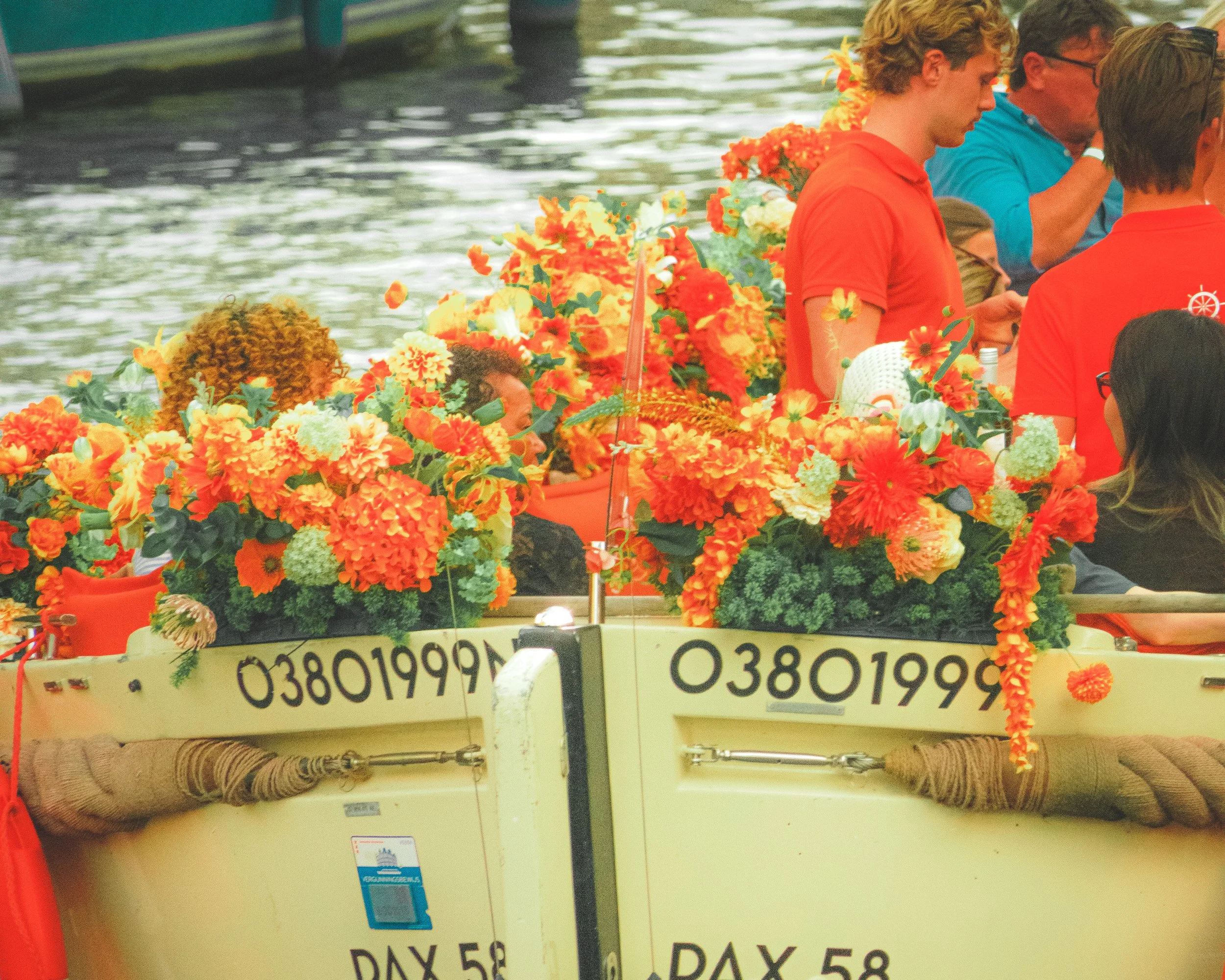 Rote und orangefarbene Blüten am Bug eines Ausflugsboots, im Hintergrund Passagiere auf dem Wasser