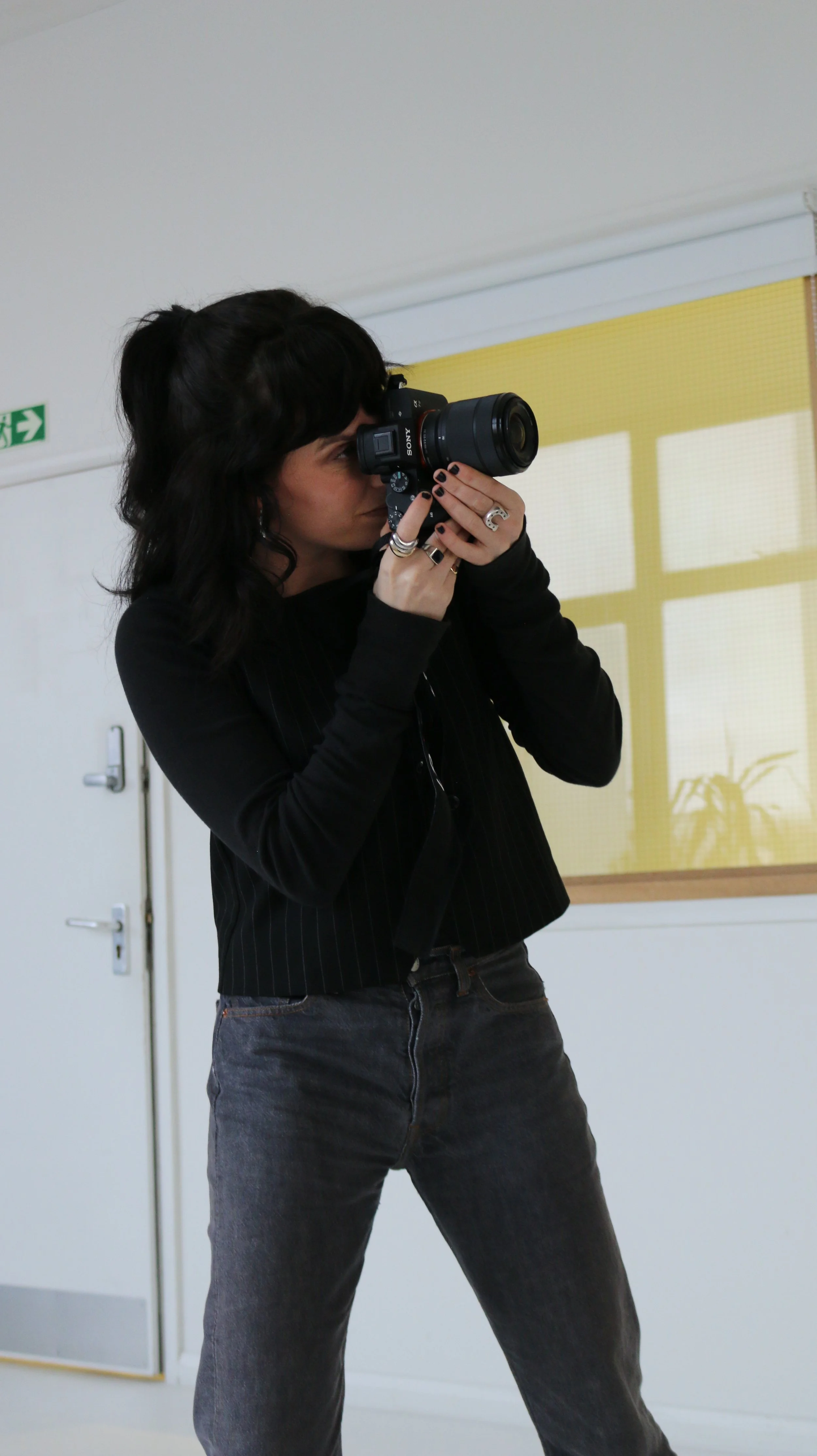 A woman with dark wavy hair, wearing a black top and dark jeans, is taking a photo with a Sony camera in a room with white walls and a yellow window shade.