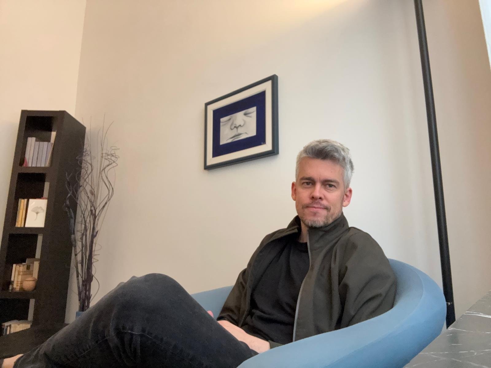A man with gray hair and a beard sitting on a light blue sofa in a modern living room, with a calm expression and a black shirt, framed artwork of a child's face hanging on the wall behind him, a tall black bookshelf with books and decor to his left, and a black floor lamp to his right.