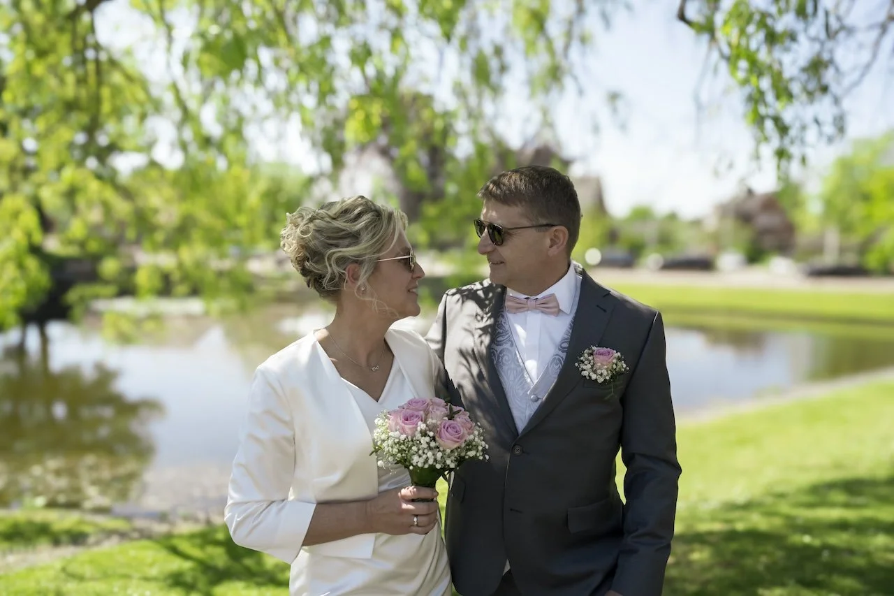 Ein Brautpaar im Freien, beide tragen Sonnenbrillen. Die Braut in weißem Kleid hält einen Blumenstrauß, der Bräutigam trägt einen grauen Anzug mit Fliege und Blumenanstecker. Im Hintergrund ist ein Teich mit grüner Umgebung.