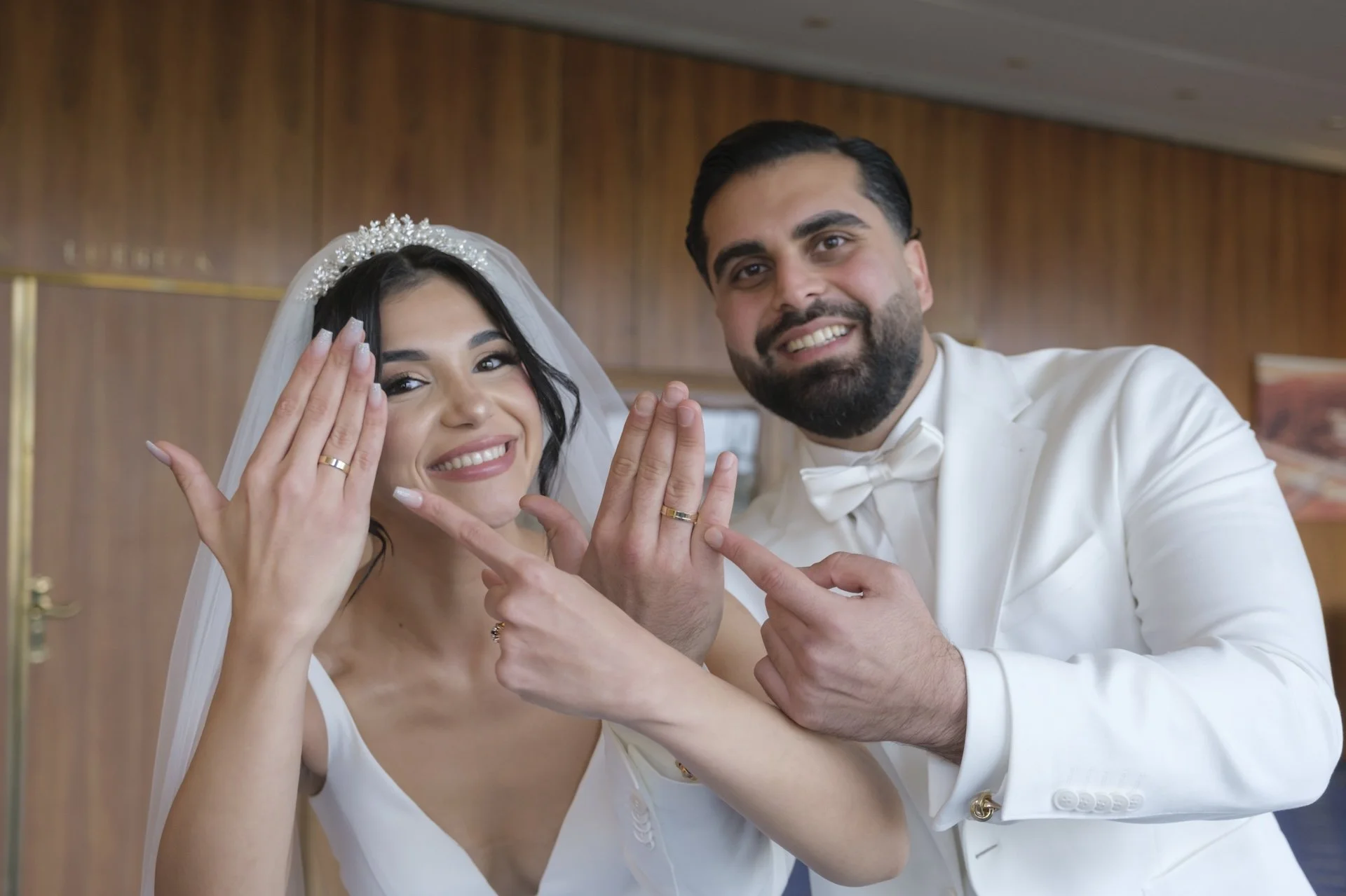 Ein Hochzeitspaar zeigt stolz ihre Eheringe in die Kamera. Beide tragen weiße Hochzeitskleidung und lächeln.