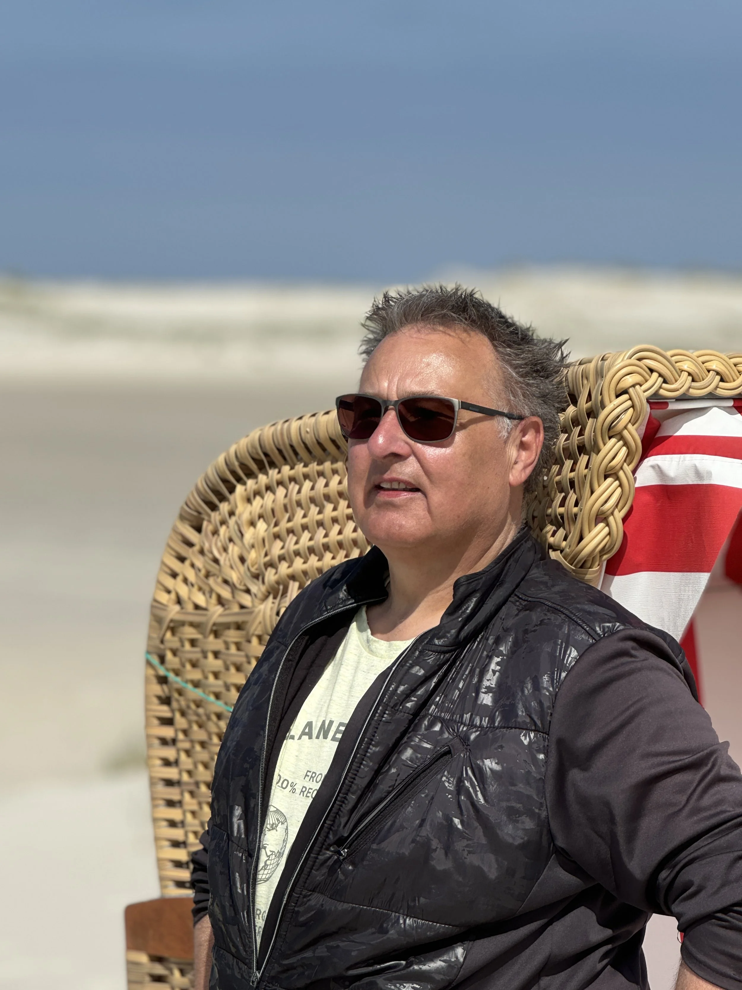 Person im Strandkorb am Strand sitzend, mit Sonnenbrille und Freizeitbekleidung. Im Hintergrund ist ein blauer Himmel sichtbar.