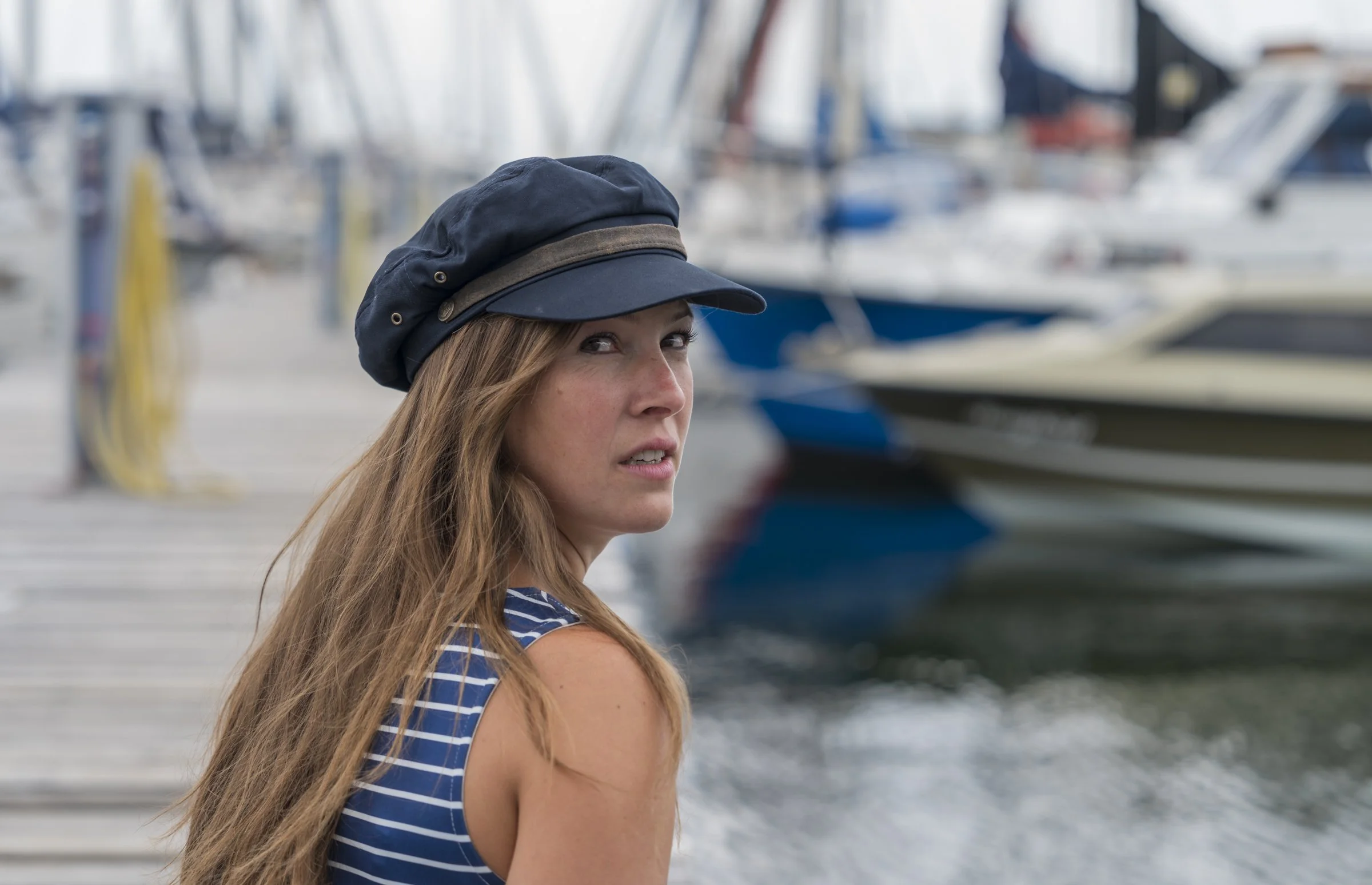 Frau mit blauer Mütze und gestreiftem Oberteil vor einem Boot im Hafen.