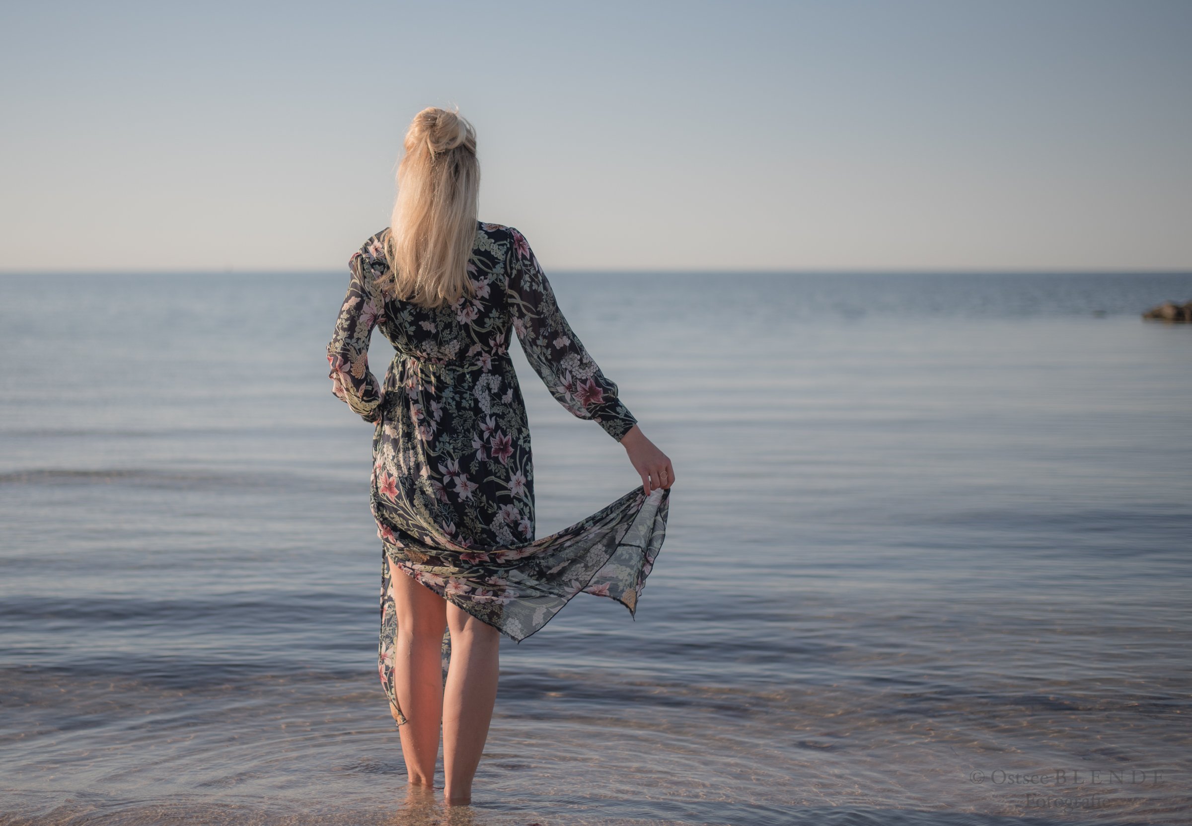 Eine Frau mit langem, blondem Haar steht im seichten Wasser am Strand. Sie trägt ein langes, geblümtes Kleid und blickt auf das Meer hinaus.