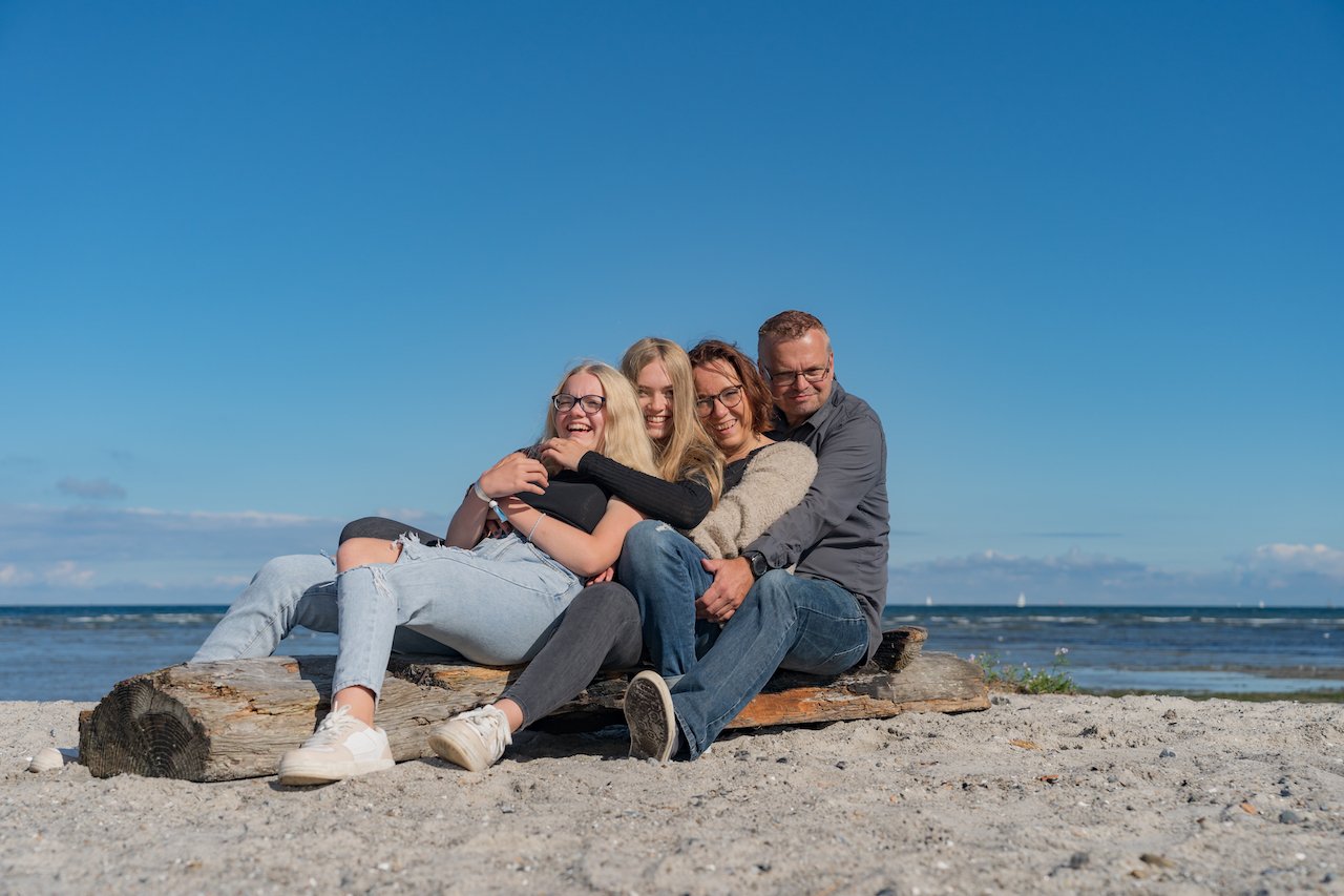 Vier Menschen sitzen lachend auf einem Holzstück am Strand vor dem Meer bei blauem Himmel.