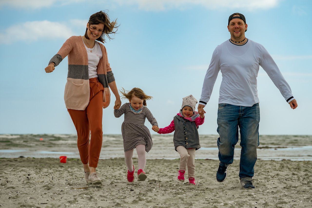 Familie, die am Strand läuft, bestehend aus zwei Erwachsenen und zwei Kindern. Im Hintergrund Meer und blauer Himmel.