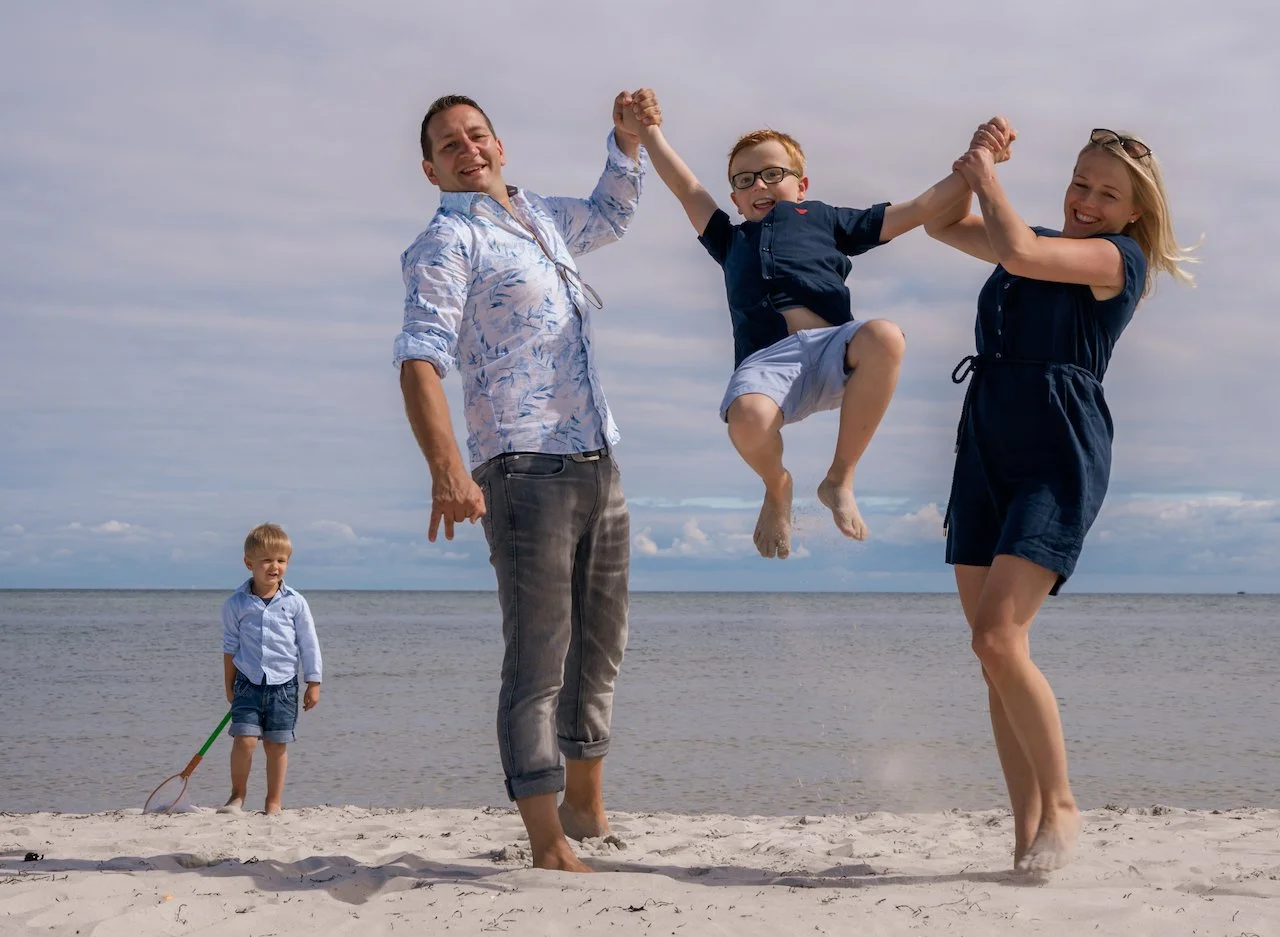 Familie am Strand, zwei Erwachsene halten ein Kind in der Luft, ein weiteres Kind mit Schläger steht im Hintergrund, Meer im Hintergrund.