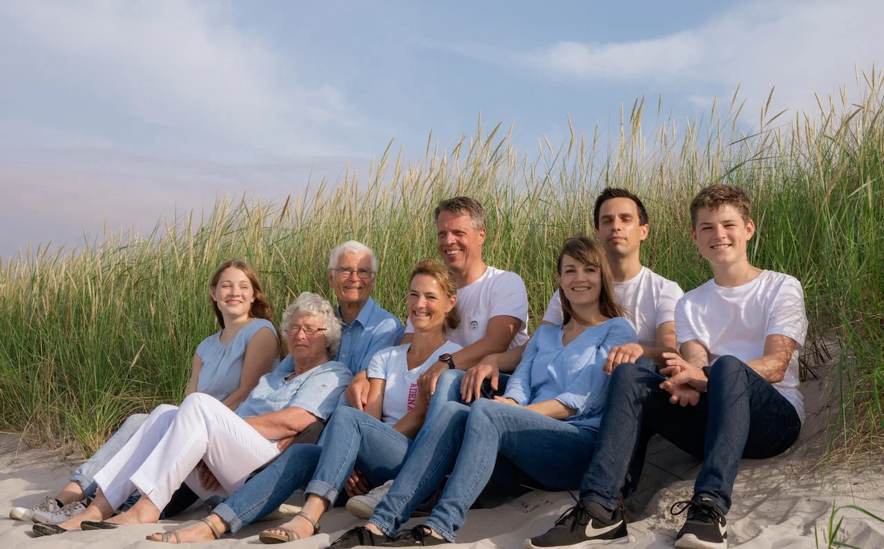 Eine Gruppe von Menschen sitzt auf einem Strand im Sand vor hohen Gräsern, lächelnd in die Kamera schauend. Der Himmel ist blau mit ein paar Wolken.