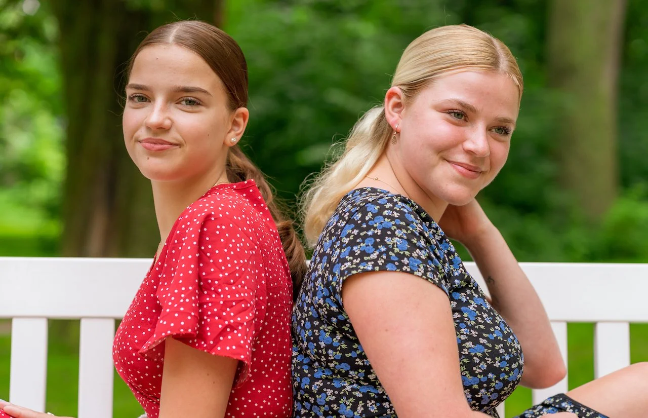 Zwei Frauen sitzen Rücken an Rücken auf einer weißen Bank im Freien, mit grüner Natur im Hintergrund. Eine trägt ein rotes gepunktetes Kleid, die andere ein blau gemustertes Kleid.