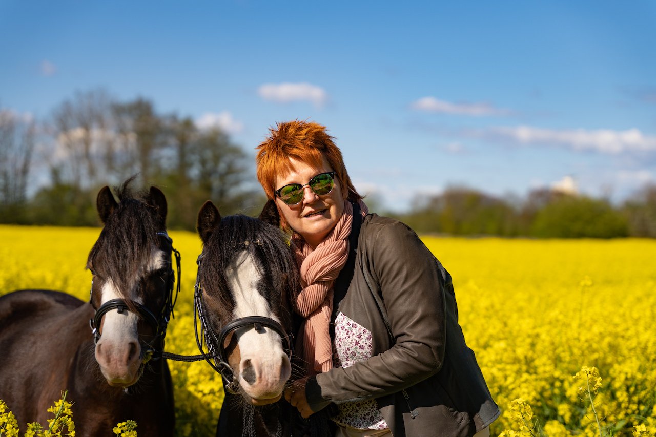 Eine Person mit orangenen Haaren steht zwischen zwei schwarzen Ponys in einem blühenden Rapsfeld unter blauem Himmel.