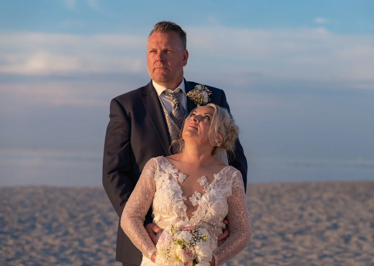 Ein Brautpaar steht am Strand, das Brautpaar trägt eine Hochzeitkleidung, die Braut hält einen Blumenstrauß, es ist Sonnenuntergang im Hintergrund.
