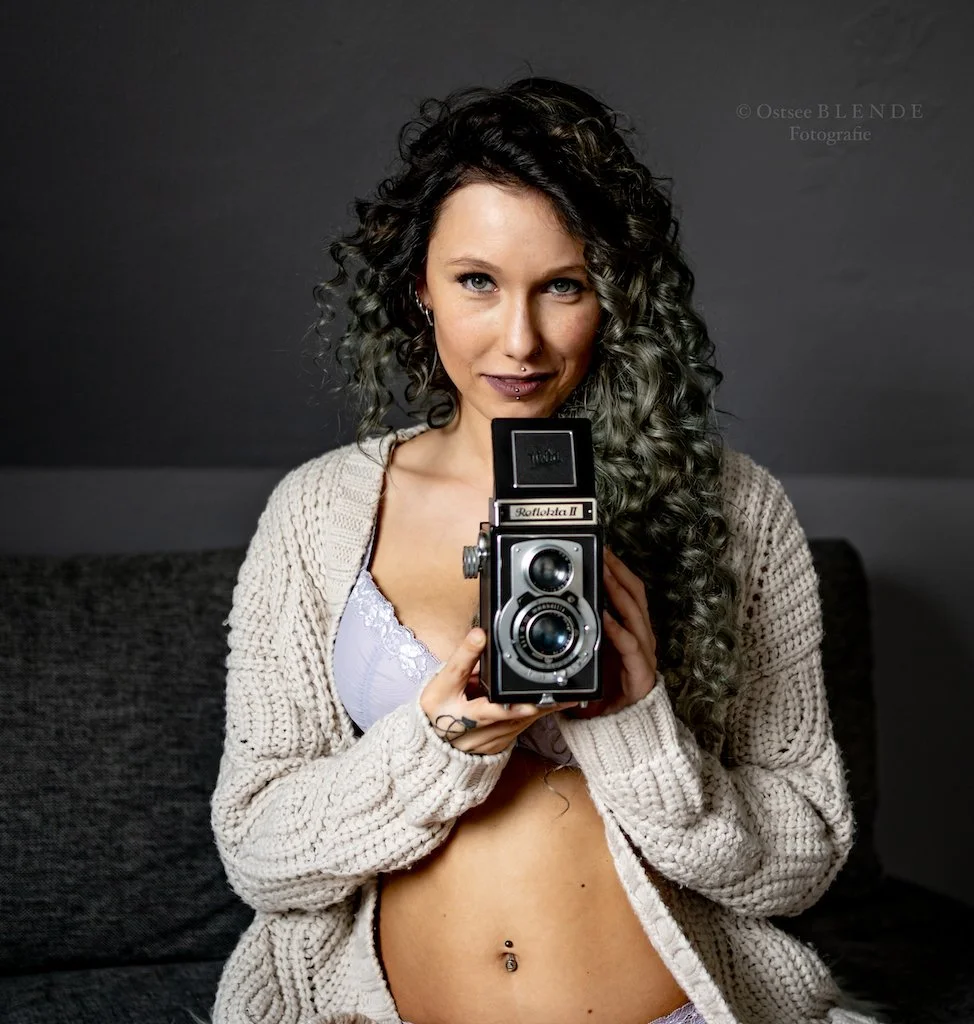 Frau mit lockigem Haar hält eine Retro-Kamera, trägt Strickjacke und BH, lächelt leicht, dunkler Hintergrund.