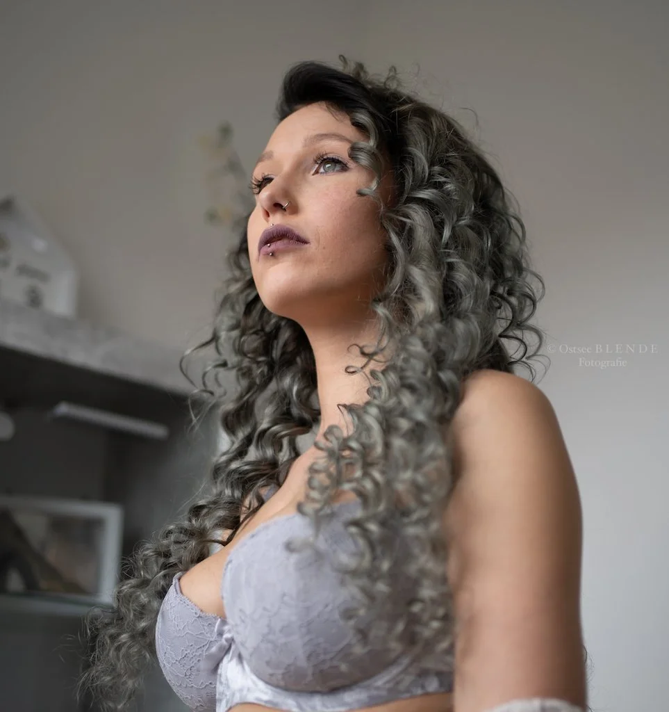 Eine Frau mit grau-gelockten Haaren trägt ein Spitzen-BH. Sie schaut nach oben.