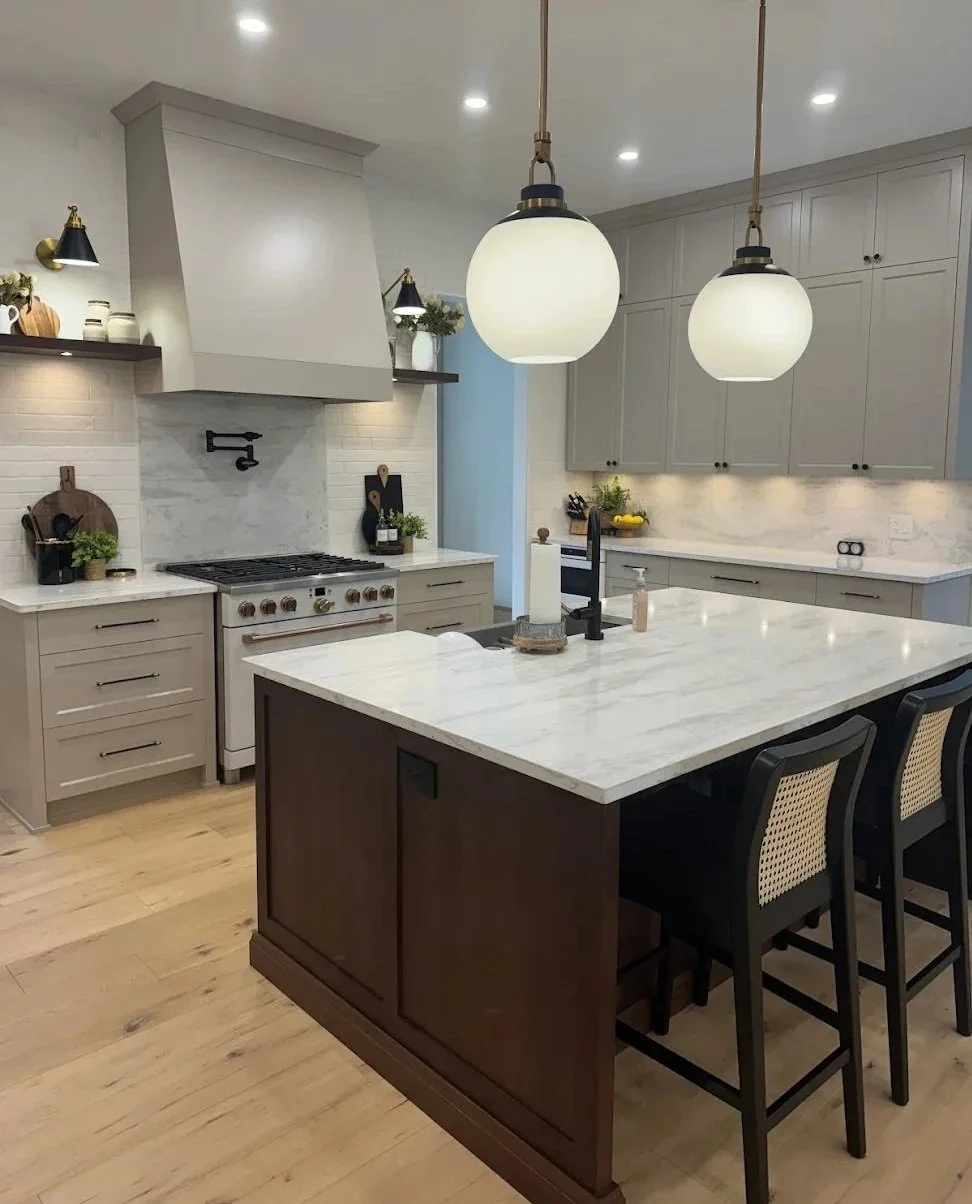 Modern kitchen with gray cabinets, white marble countertops, a kitchen island with a dark wood base, black chairs, two large pendant lights, and various decorative items.