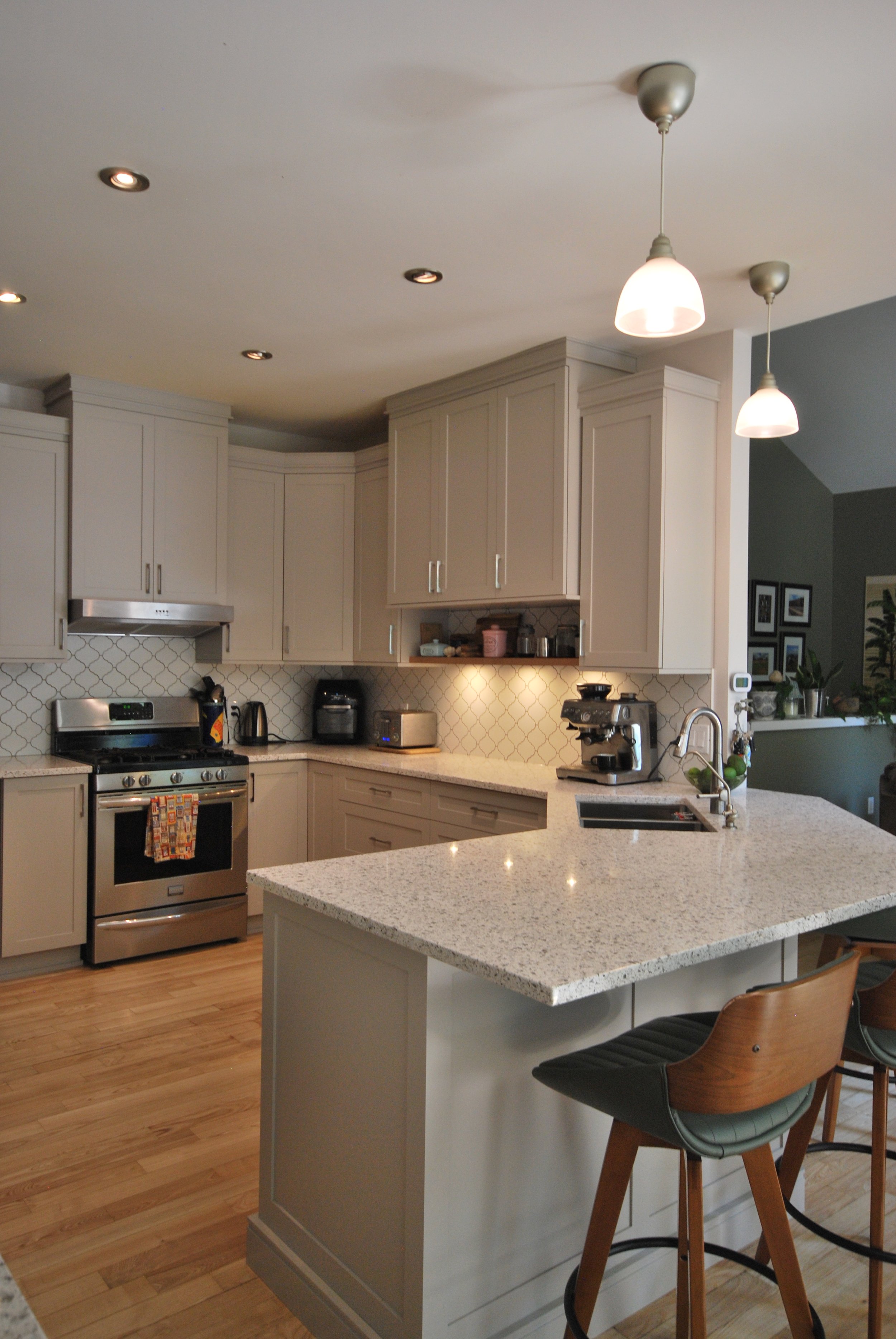 A modern kitchen with white cabinets, stainless steel appliances, a kitchen island with a white countertop, pendant lights, and wooden stools. The kitchen has wooden flooring and various small appliances on the counter.