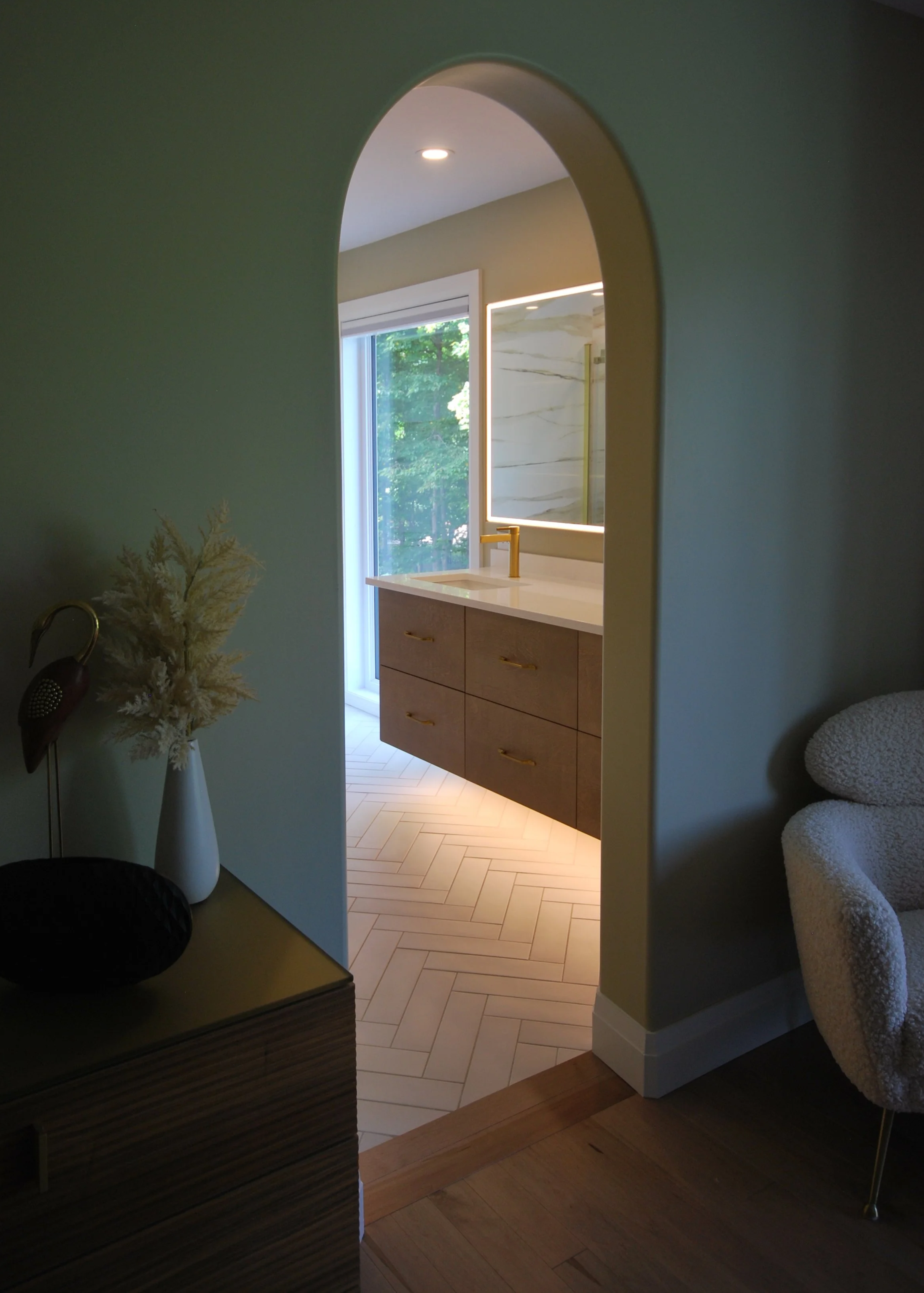 A view through an arched doorway into a bathroom with a vanity, marble wall, and large window showing greenery outside.