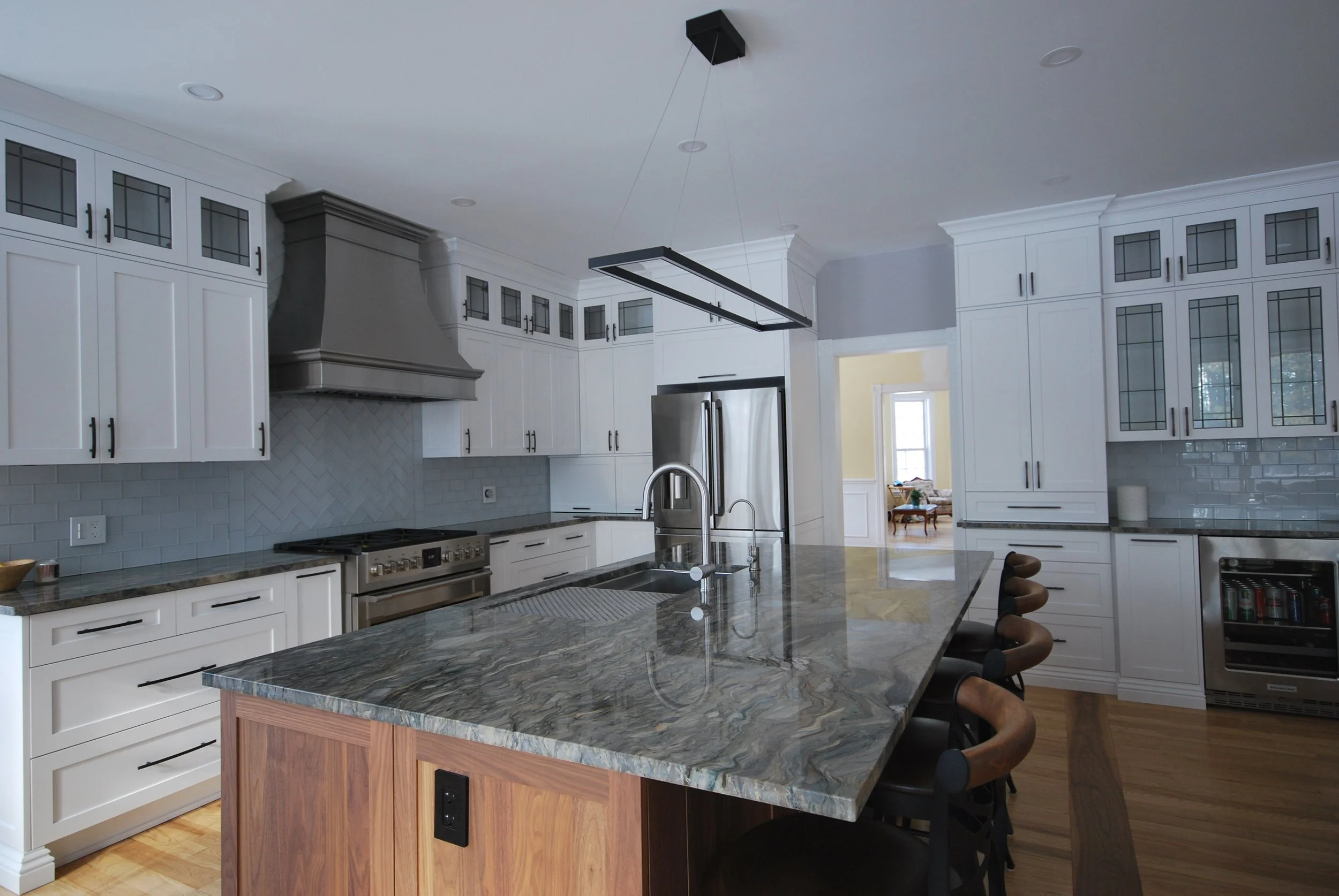 Modern kitchen with white cabinets, a large marble island, stainless steel appliances, and a gray range hood. Black barstools are lined up at the island, and a doorway leads to a dining area with natural light.