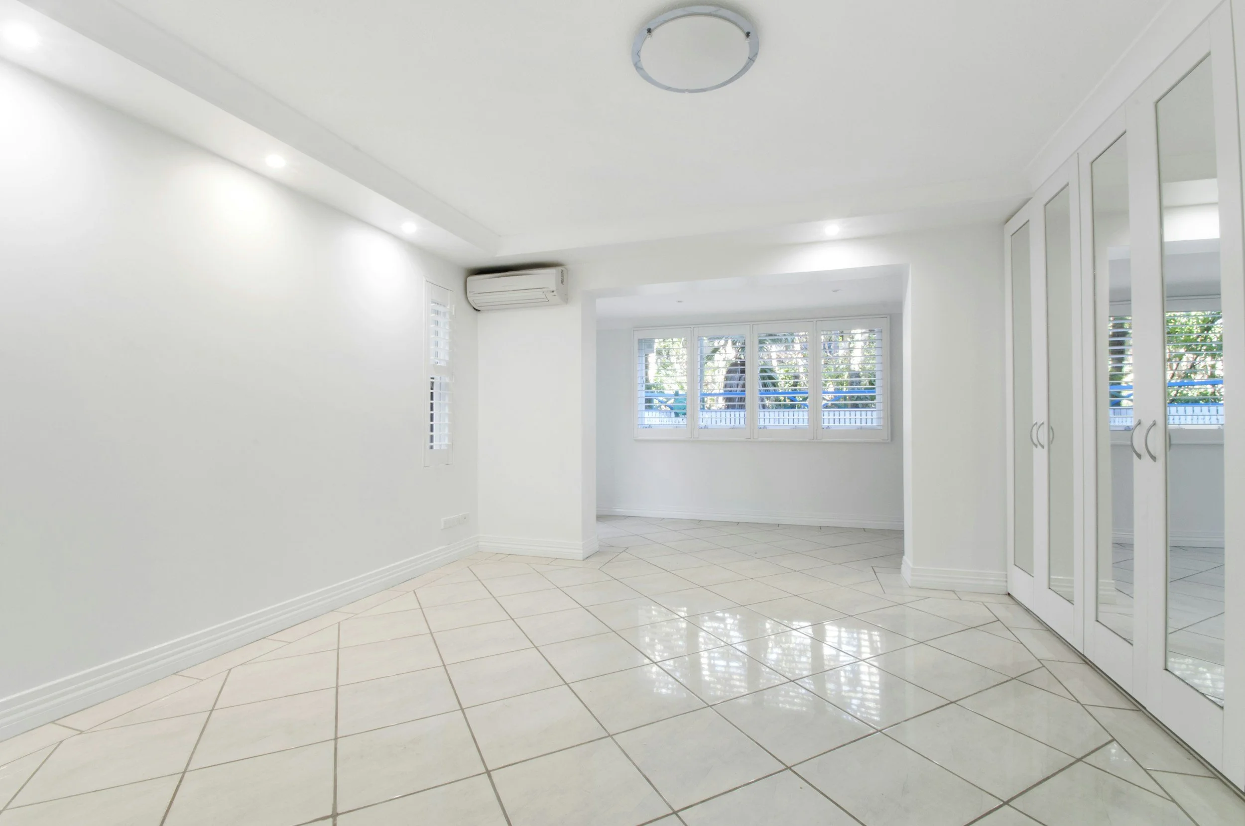 Empty white room with large windows, tiled floor, built-in shelves, and air conditioning unit on the wall.