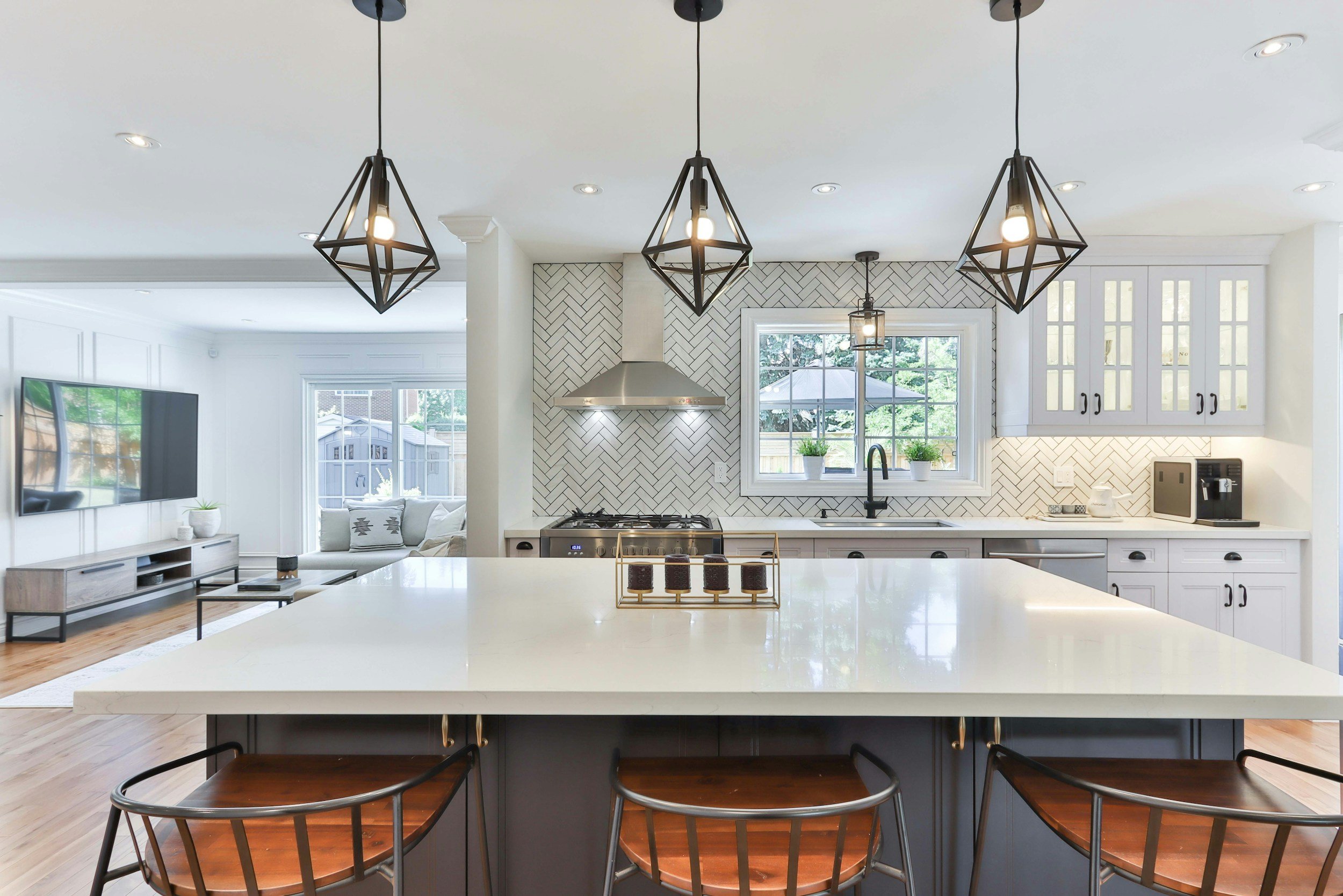 Modern kitchen with three black geometric pendant lights hanging over a white island, subway tile backsplash, stainless steel range hood, large window with two potted plants, white cabinetry with black handles, and open living room with TV on the wall and a beige sofa.