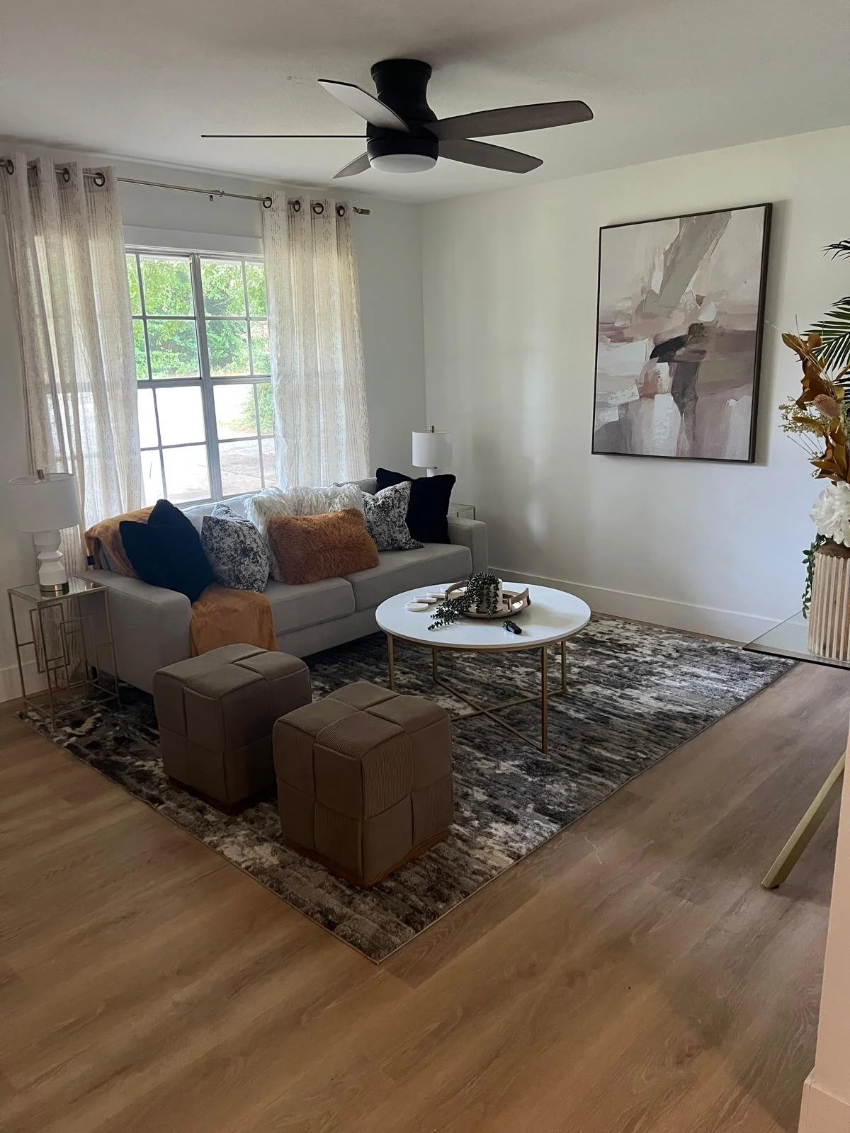 A living room with a white sofa adorned with various blue, white, and tan throw pillows, a round white coffee table with decorations, two beige ottomans, a large abstract painting on the wall, a window with sheer curtains, a potted plant, and a black ceiling fan.