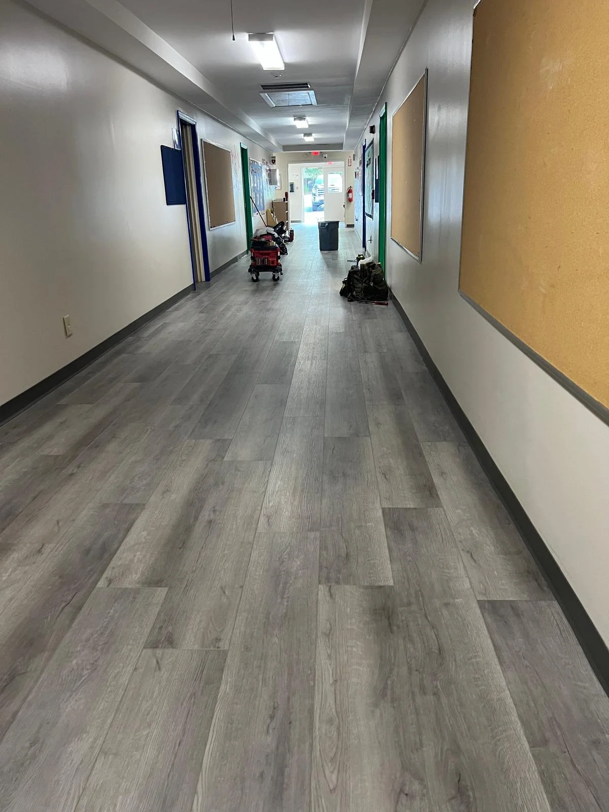 Empty hallway in a school or office building with bulletin boards on the walls, cleaning supplies, and a trash bin near the entrance at the end of the hall.