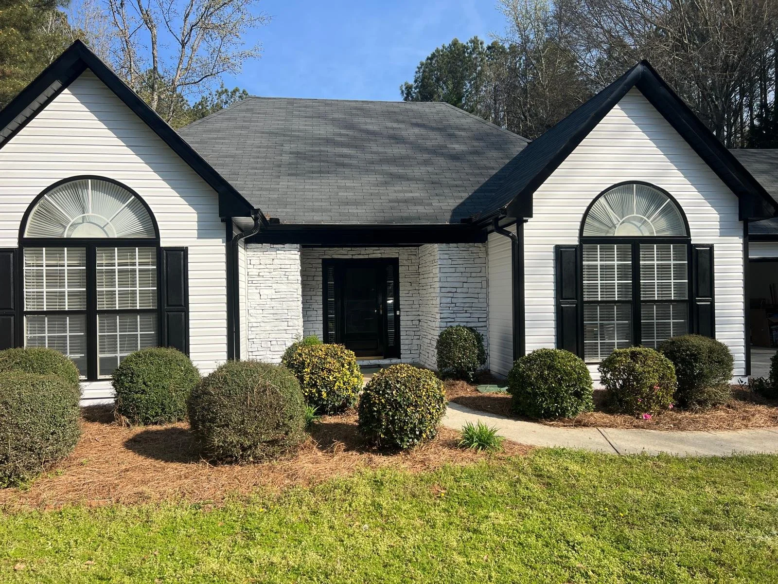 Front view of a house with white siding, black shutters, and large arched windows. There are bushes and a small lawn in front of the house.