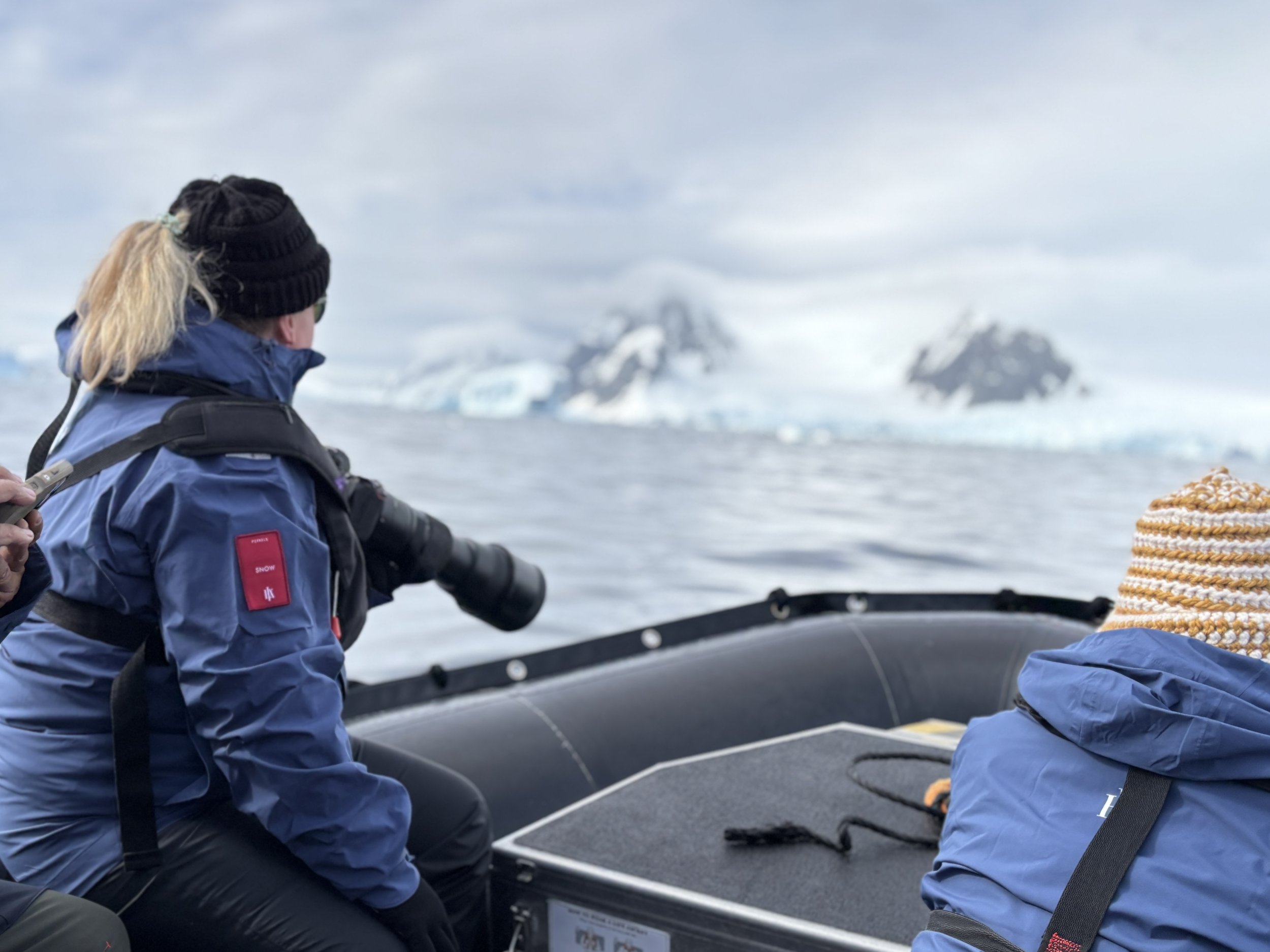 A person wearing a black knit hat, sunglasses, and a blue jacket with a red patch, sitting on a boat and looking at snowy mountains and ice in the distance.