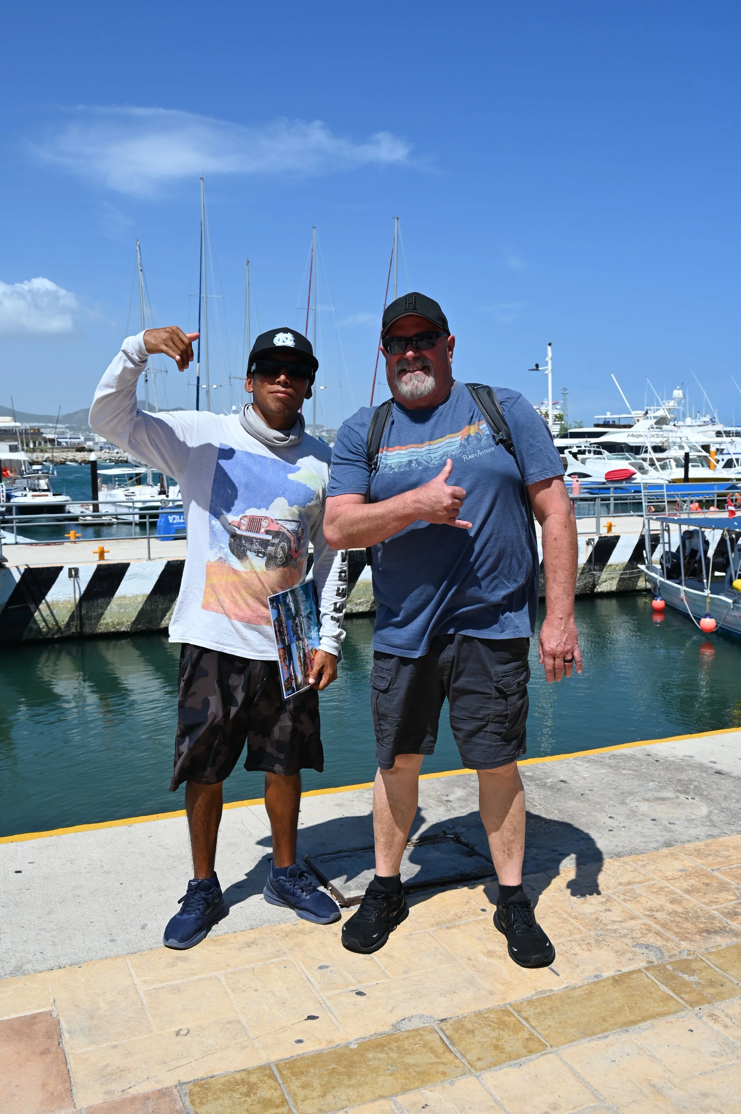 Two men standing near a marina filled with sailboats. The man on the left is wearing sunglasses, a black cap, a white long-sleeve shirt with a graphic print, and shorts. The man on the right is wearing sunglasses, a black cap, a blue t-shirt, and shorts, giving a thumbs-up. The sky is blue with some clouds, and the water is calm.