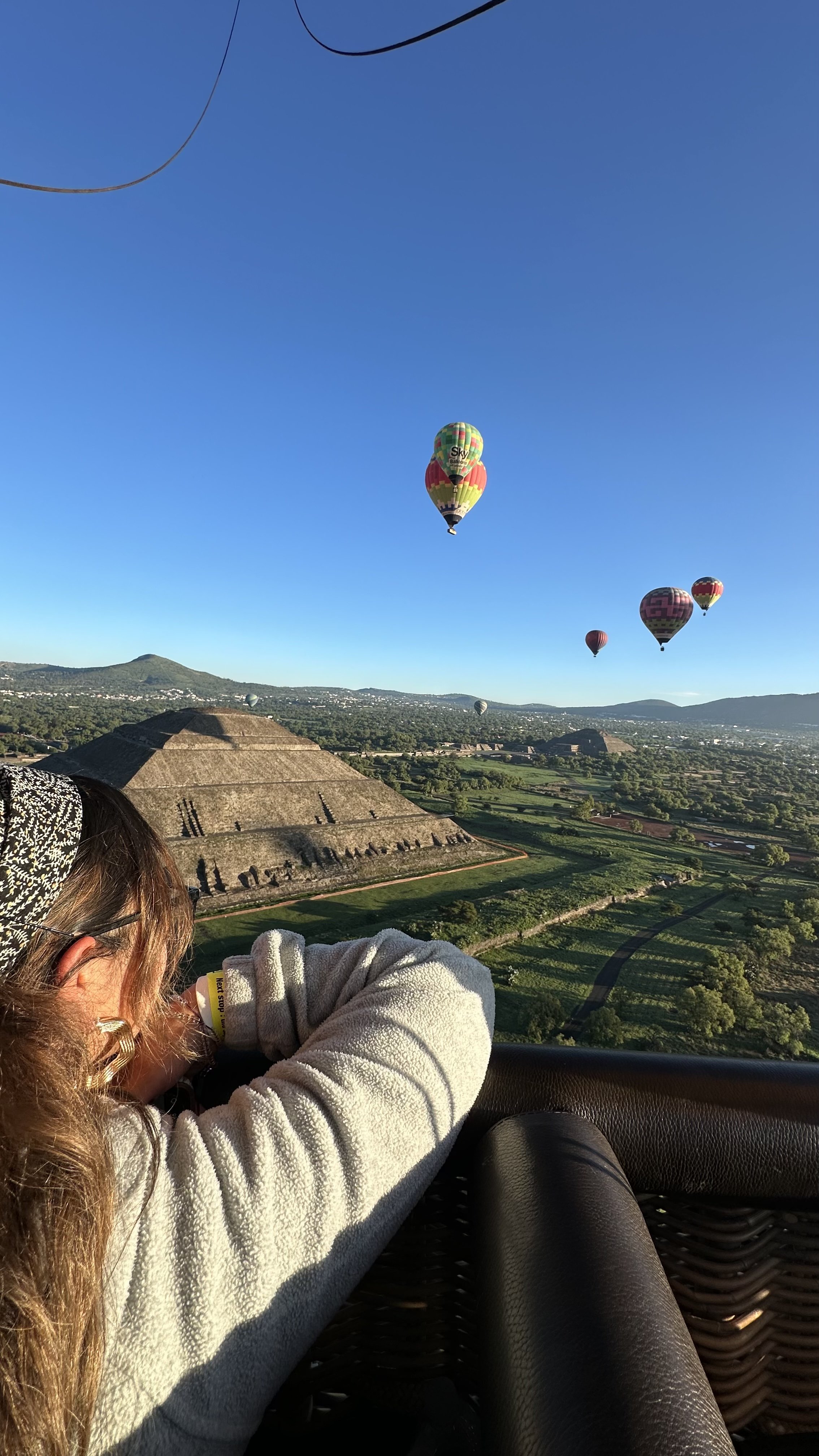 A woman lying in a hot air balloon basket taking a photo of multiple hot air balloons in the sky over a landscape with pyramids and greenery.
